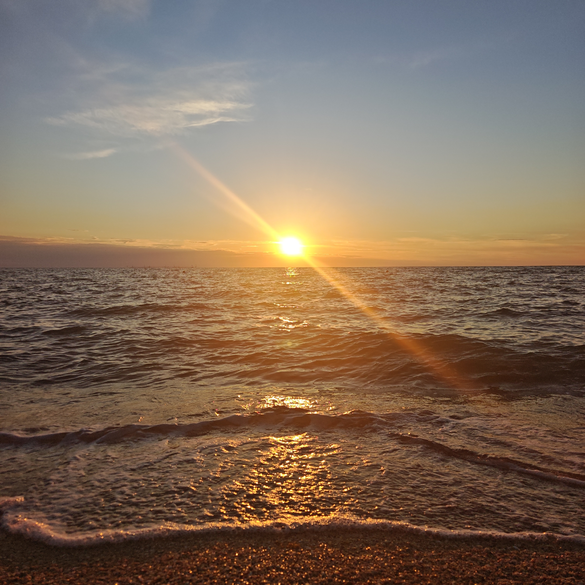 The image depicts a serene seascape at sunset. The sun, a bright, luminous orb, is positioned slightly left of center on the horizon, casting a warm, golden glow across the water and sky. Sunbeams radiate outwards from the sun, creating streaks of light that reflect brilliantly on the rippling surface of the sea. Small, gentle waves are breaking on a pebbly shore in the foreground, with frothy white foam tracing the edge of the water. In the distance, along the horizon line, faint structures resembling poles or buoys are visible. The sky transitions from a pale blue at the top to a warm, hazy orange near the horizon, with wispy clouds scattered across its expanse. The overall impression is one of tranquility and natural beauty during the golden hour. The location is identified as Chelyadinovske rural settlement, Ukraine.