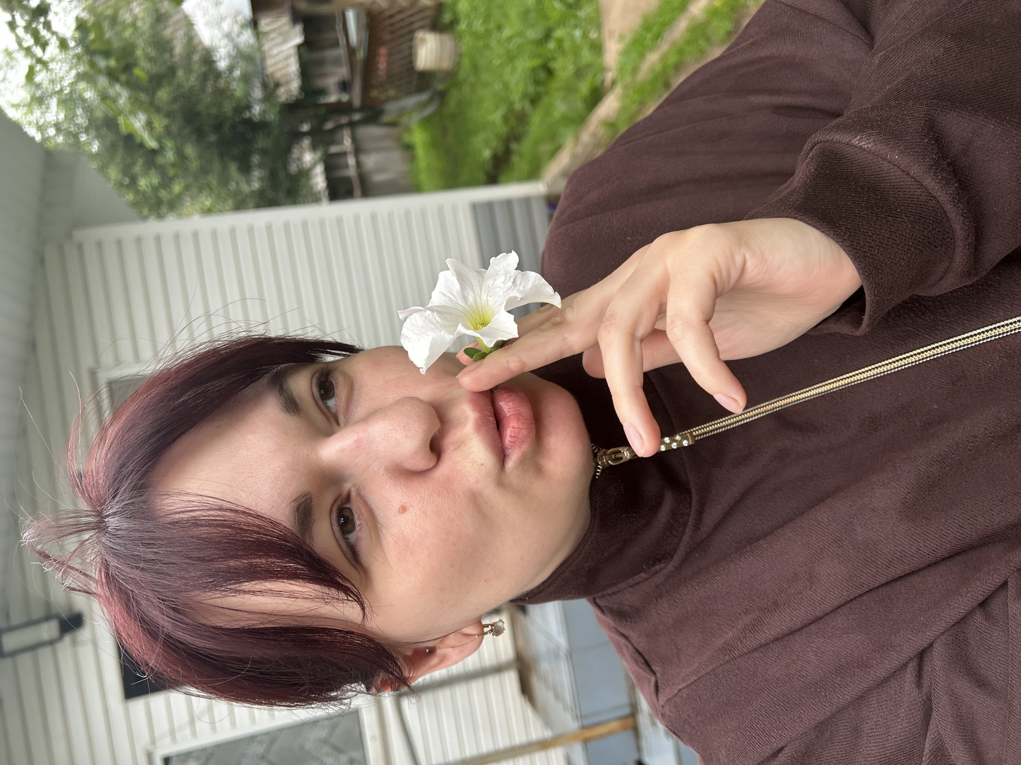 A young woman with short, dark red hair, wearing a brown zip-up jacket, holds a white flower to her lips with her right hand. She is outdoors in a semi-urban or rural setting, with a white-sided building with horizontal siding in the background, and a blurred green, grassy area with trees and a wooden fence to the right. The woman is facing to her right, with her lips pursed as if to kiss the flower. She has a small earring in her left ear. The lighting suggests it is daytime, possibly overcast. The location is identified as Fokino, Russia.