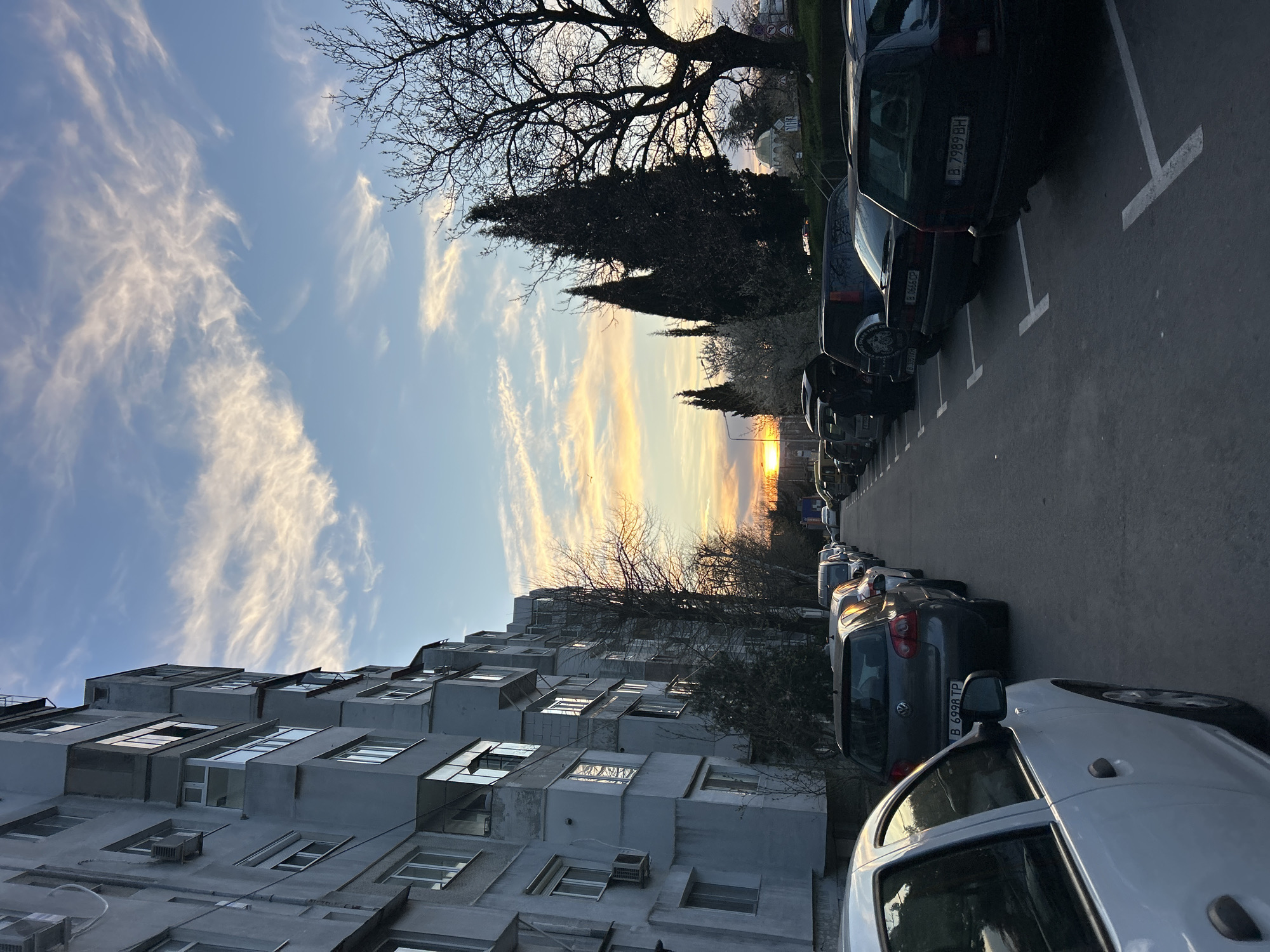 The image depicts a street scene at sunset in Varna, Bulgaria. On the right, cars are parked in designated parking spaces along the roadside. Vehicle license plates are visible on some of the vehicles. The street extends into the distance, with additional parked cars visible further down. A multi-story concrete apartment building is on the left side of the frame. The sky above features light clouds, with a sun setting on the horizon. Silhouetted trees are also visible between the building and the cars.