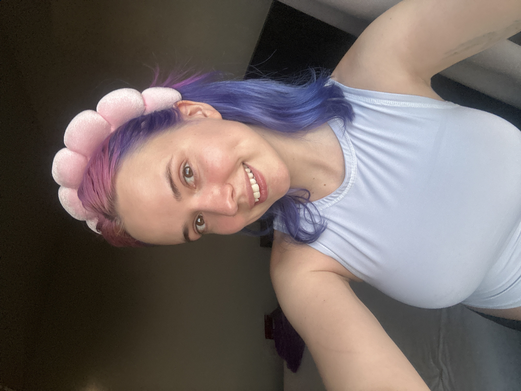 A young woman is posing for a selfie. She is wearing a light blue tank top and a pink flower headband. Her hair is dyed with purple and pink streaks. She is smiling and looking at the camera. The background is a dark room. There is a window to the right of the woman. There is no verifiable information to place this image in Ramonville-Saint-Agne, France.