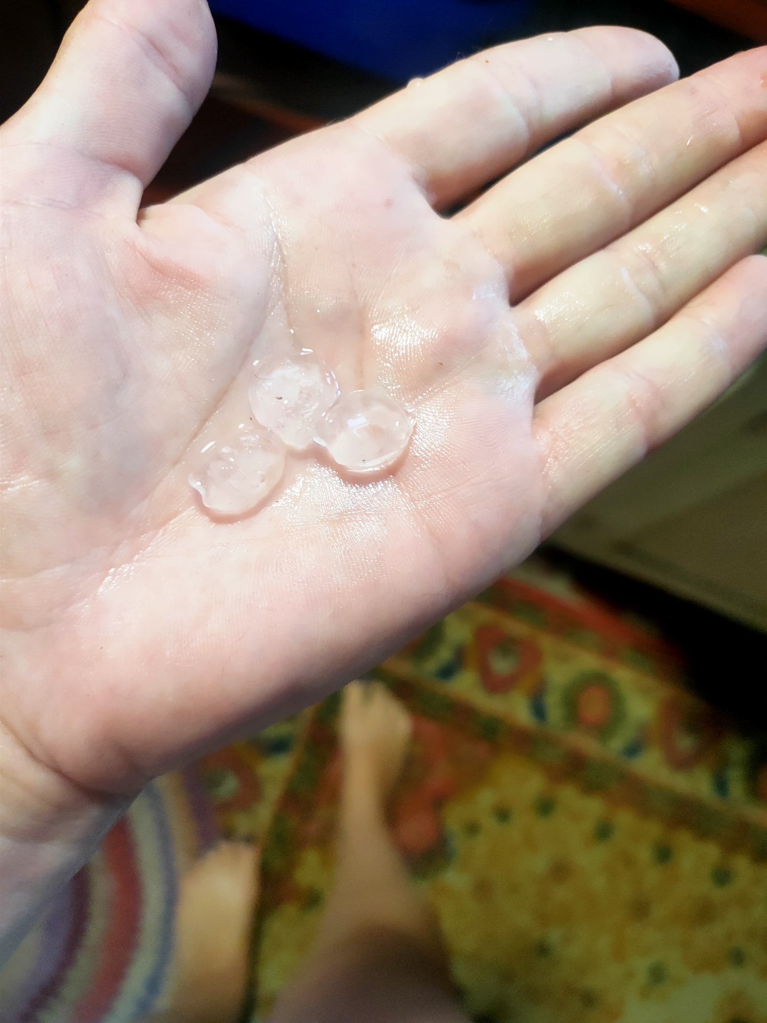A close-up shot shows a person's palm holding three small, translucent, spherical objects that appear to be hail. The palm is slightly wet, and the skin is pinkish. The background is out of focus, but a colorful rug and a person's leg are visible. The image was taken in Kremenchuk, Ukraine.