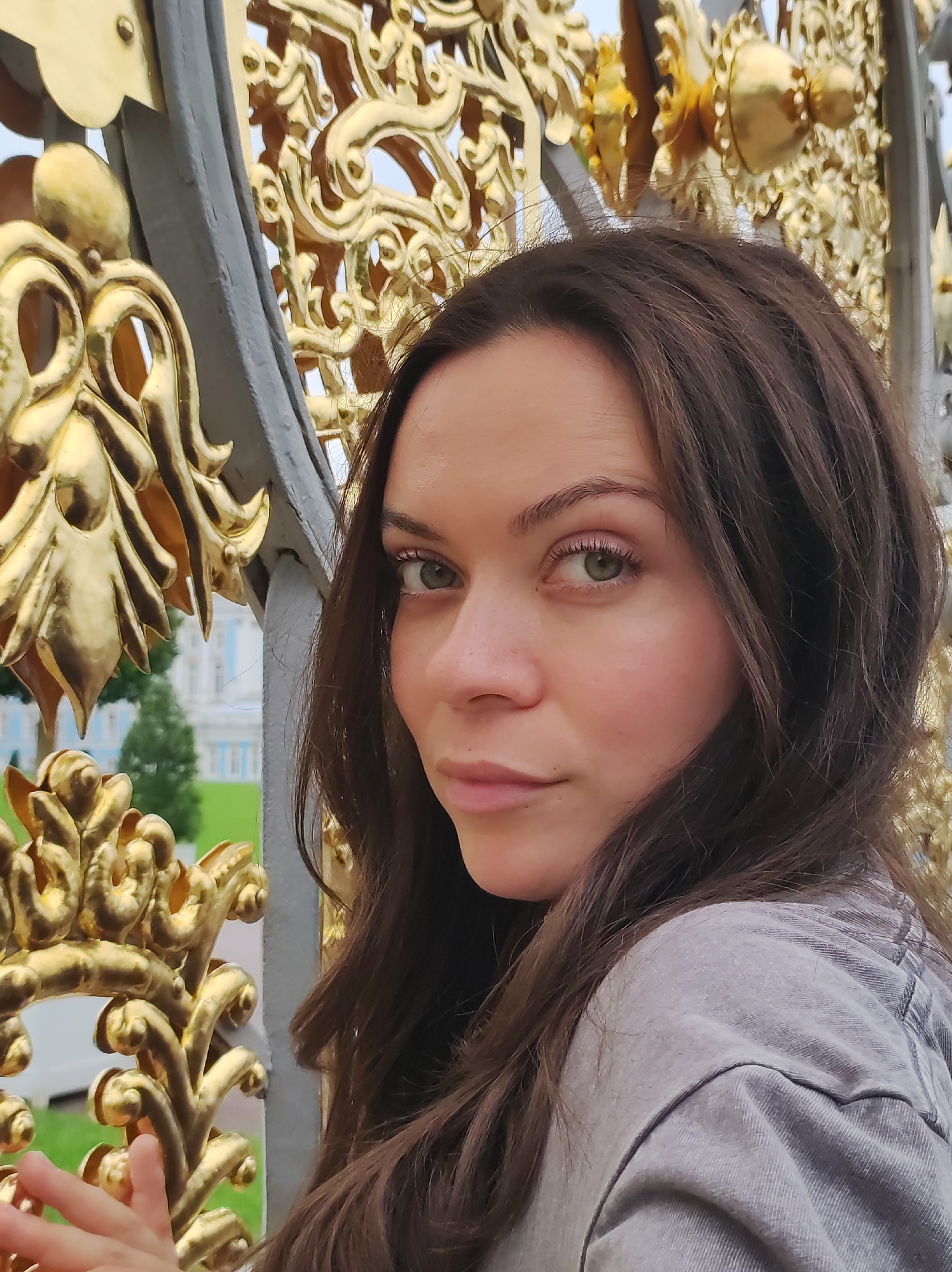 The image shows a close-up portrait of a young woman with long, dark brown hair and green eyes. She is looking slightly to her left, with her head tilted. She is wearing a grey shirt. Behind her, a decorative gold gate with intricate patterns is visible. In the background, a building with white walls and blue trim, along with green trees, can be seen, suggesting a park or garden setting. This appears to be a view from the Catherine Palace in Pushkin, Russia, specifically from its ornamental gates.
