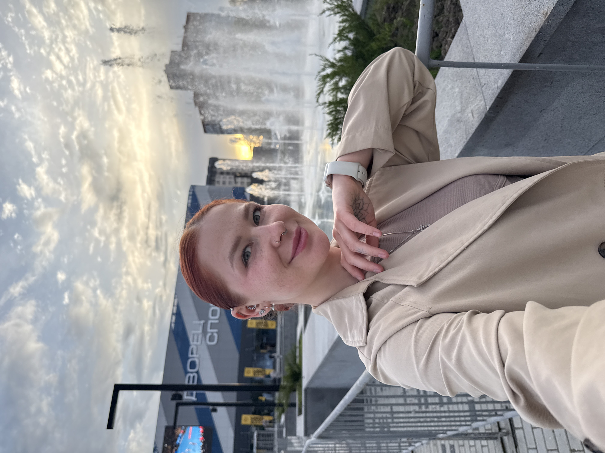 A medium shot depicts a fair-skinned woman with freckles, red hair pulled back, wearing a beige trench coat, a matching top, and multiple earrings. She has a nose ring and a small tattoo on her hand, resting her hand near her neck. Behind her is a fountain, a building with the word "Дворец Спорта" (Palace of Sports) in Cyrillic script, and a cloudy sky. The location is Rostov-on-Don, Russia.
