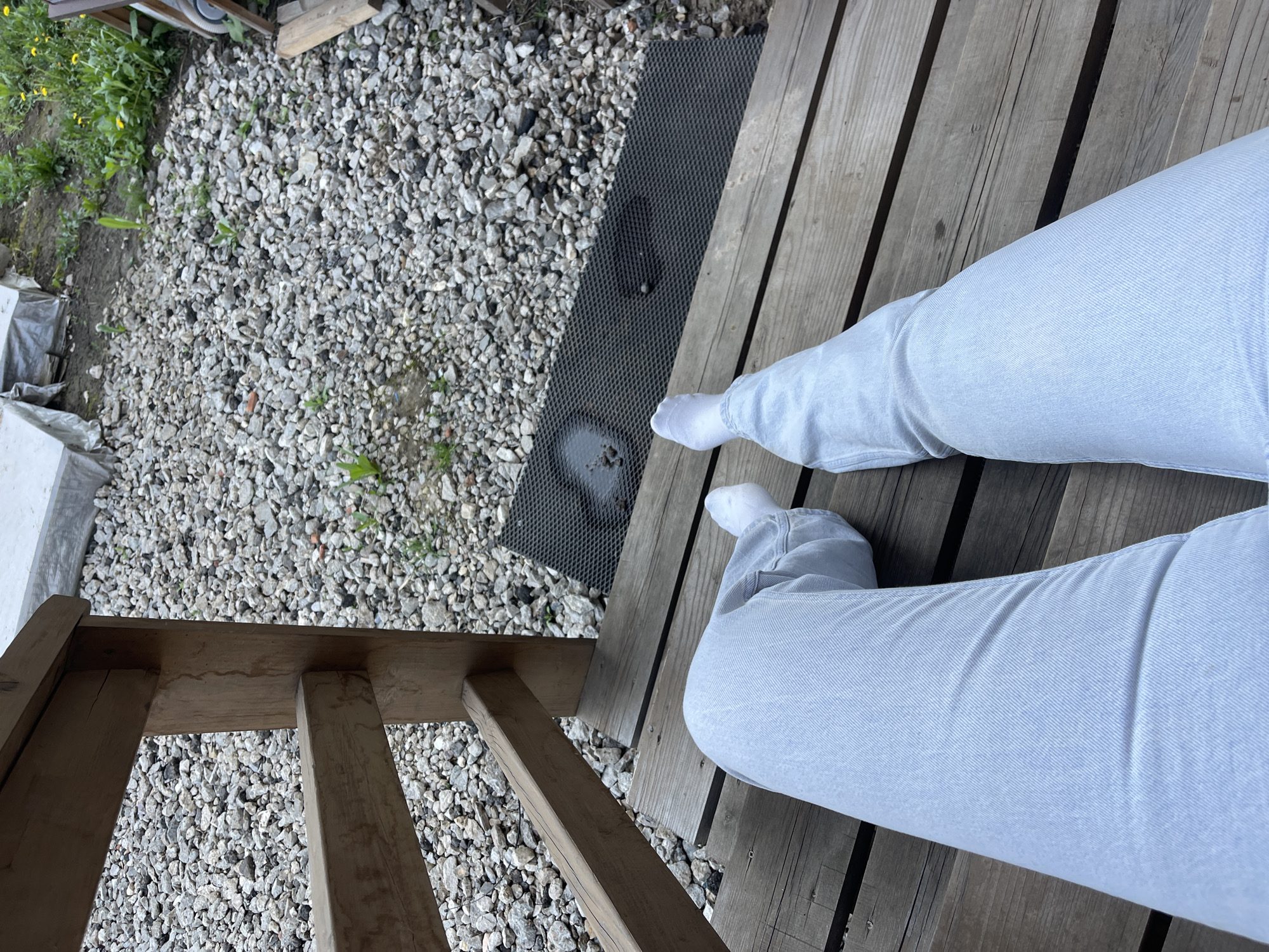 A person wearing light blue jeans and white socks is sitting on a wooden deck. To the left, a black mat sits next to a gravel area with some greenery. A wooden railing is visible on the left side of the frame. The location appears to be outdoors. There is no evidence to suggest this is in the city of Мосметаллоконструкция Онт, Russia.