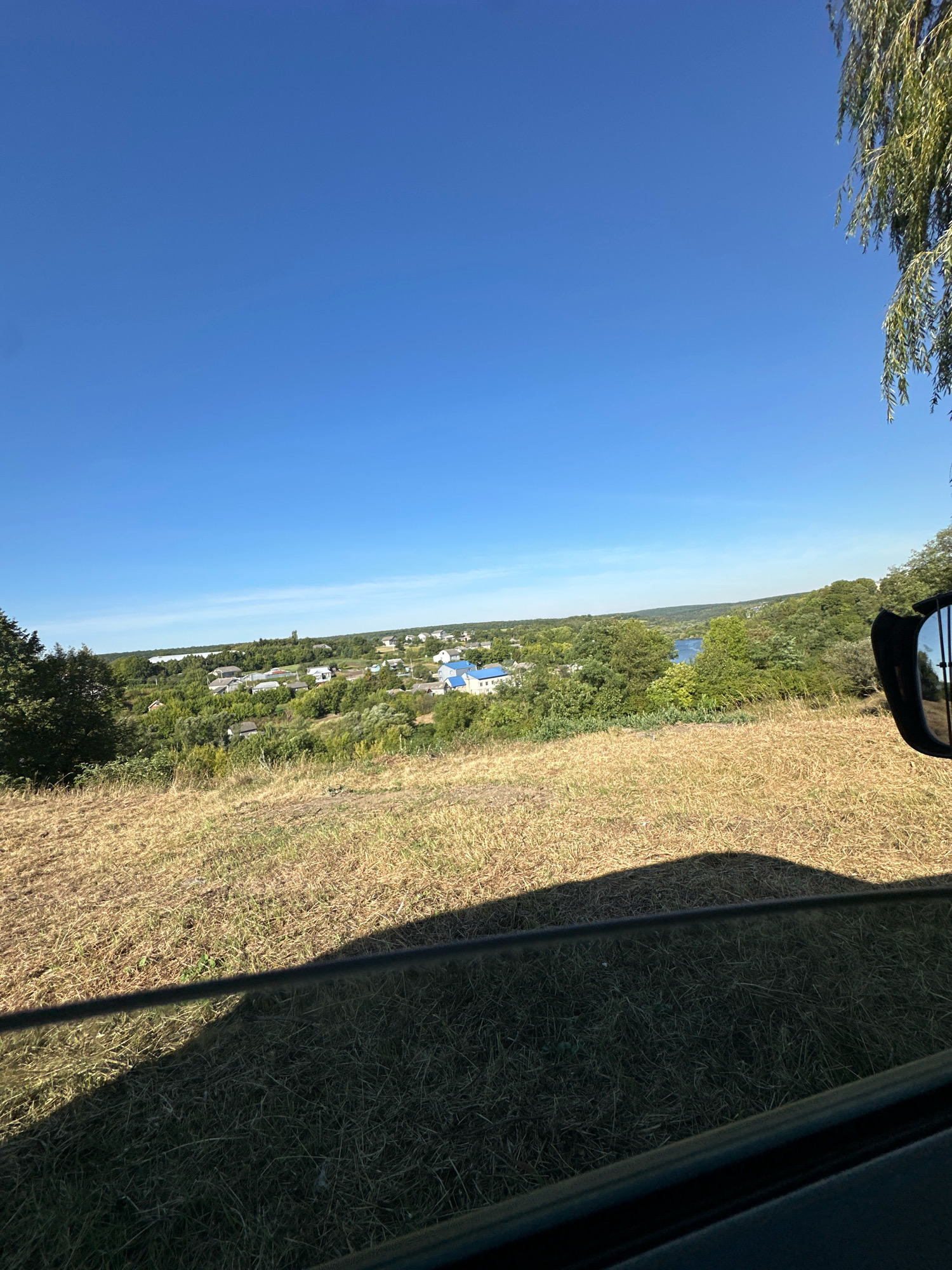 This is an eye-level, exterior, long shot of a rural landscape in Makiv, Ukraine. The foreground is dominated by dry, golden grass, casting a dark shadow on the ground from an unseen object. A dark, reflective bar runs horizontally across the image, potentially the edge of a car window. In the mid-ground, a small village is visible with houses featuring blue roofs, nestled amongst lush green trees. Beyond the village, a body of water, likely a river, reflects the clear blue sky. The background consists of rolling hills covered in dense foliage, under a vast, cloudless blue sky. A branch of a weeping willow tree hangs into the frame from the upper right corner. The scene appears to be captured on a sunny day.