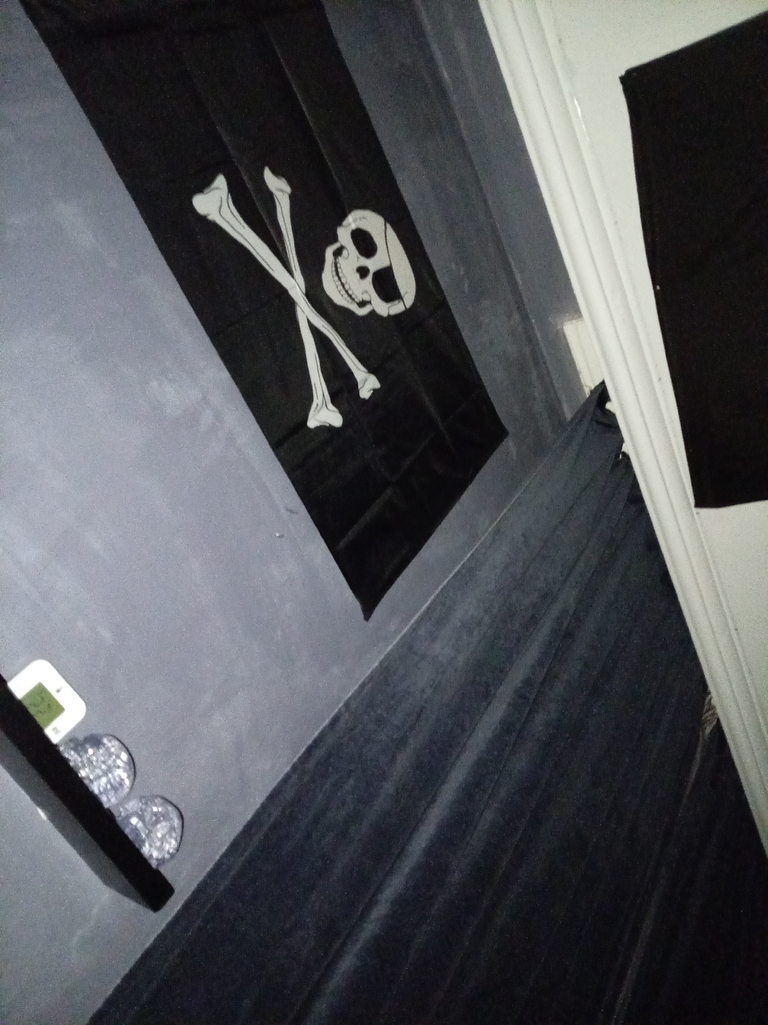 A photograph taken indoors at an oblique angle shows a black Jolly Roger flag hanging against a gray wall. A small black shelf to the left displays a small white electronic device and two glass orbs. To the right of the flag, dark gray or black curtains drape down. A white door frame is visible at the right edge of the frame, with a black object partially obscuring the right corner of the door. The location is not identified.