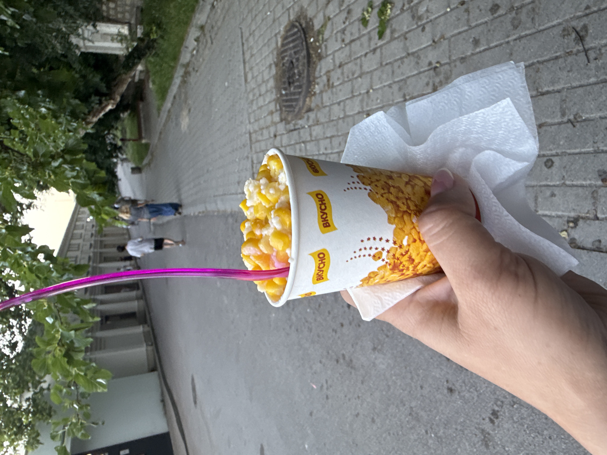 A close-up shot shows a hand holding a paper cup filled with corn and a pink straw. The cup has "ВКУСНО" printed on it in yellow lettering, along with a pattern of corn kernels. The hand is wrapped in a white paper napkin. In the background, a paved walkway with a manhole cover is visible, with people walking in the distance. Trees and buildings line the walkway, suggesting an outdoor urban setting.