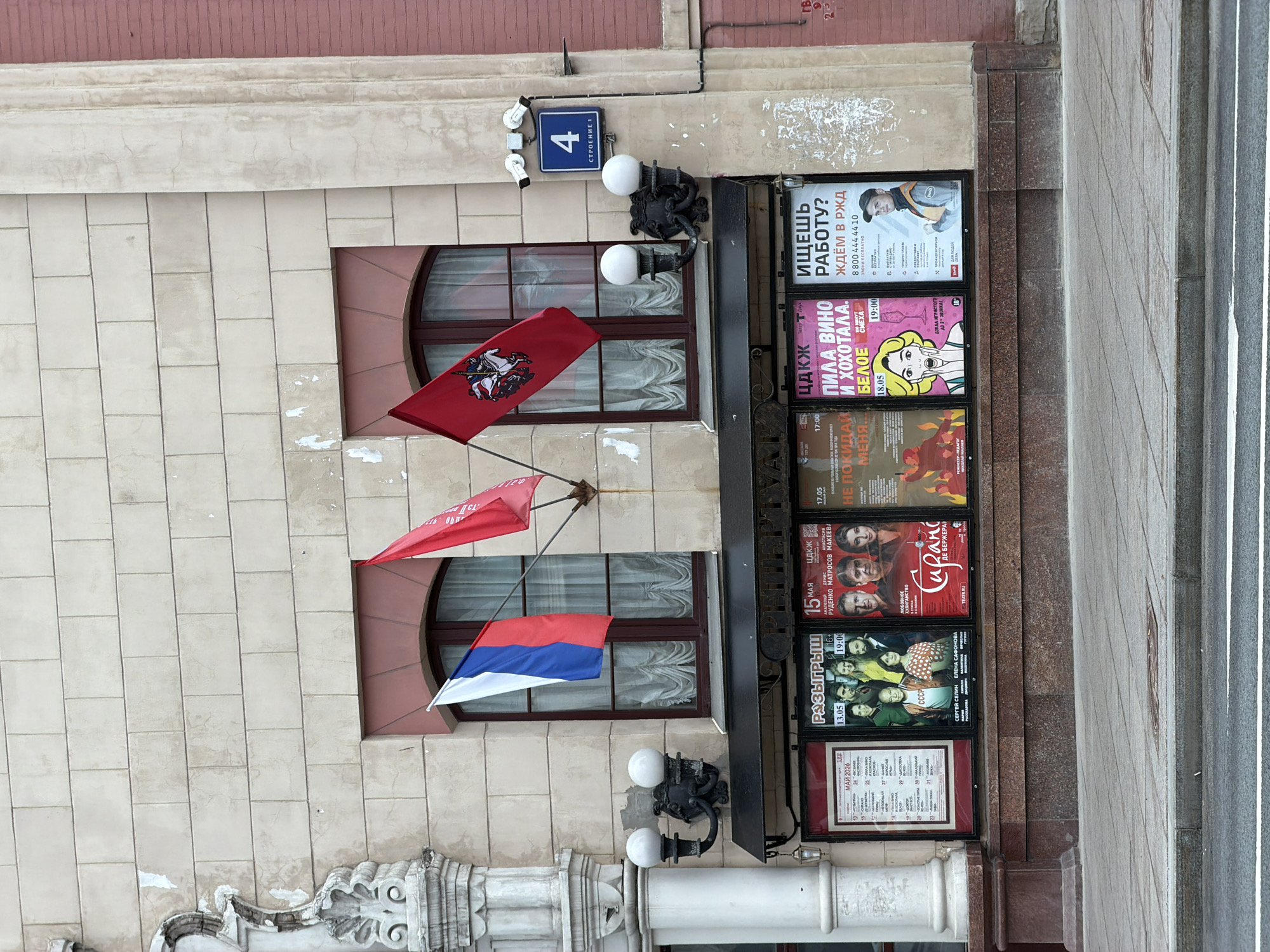 Here's a detailed description of the image:
**Scene:** An exterior shot of a building facade in Moscow, Russia. The building appears to be a theater or cultural center, indicated by the posters displayed near the entrance.
**Architecture:** The building is constructed with light-colored, rectangular stones. It has two windows with arched tops and dark brown frames. Architectural detailing includes decorative scrollwork at the base.
**Flags:** Three flags are mounted on poles attached to the wall between and above the windows. From top to bottom, the flags appear to be:
1. A red flag bearing an emblem, likely associated with Moscow.
2. A plain red flag.
3. The flag of Serbia (red, blue, and white horizontal stripes).
**Posters:** A series of posters hang in frames to the right of the windows. The posters appear to advertise theatrical performances or other events. The posters have text in Russian and include actors' faces, titles, and dates.
**Street Number:** A blue square sign with the number "4" and some additional text in Russian indicates the building's address.
**Lighting:** Decorative exterior lights are mounted on either side of the windows and near the entrance. The lights have spherical white globes.
**Street:** Adjacent to the building is a sidewalk made of grey stone tiles.
**Signs:** Advertisements that say "Ищешь работу? Ждём в РЖД" which translates to "Looking for work? We are waiting for you at Russian Railways" are hanging at the top of the poster display.
**Overall Impression:** The scene suggests a cultural venue in a historic part of Moscow. The flags and posters provide contextual information about the building's function and location. The time of day is unclear, but the presence of the lights suggests it might be either early morning or late afternoon.