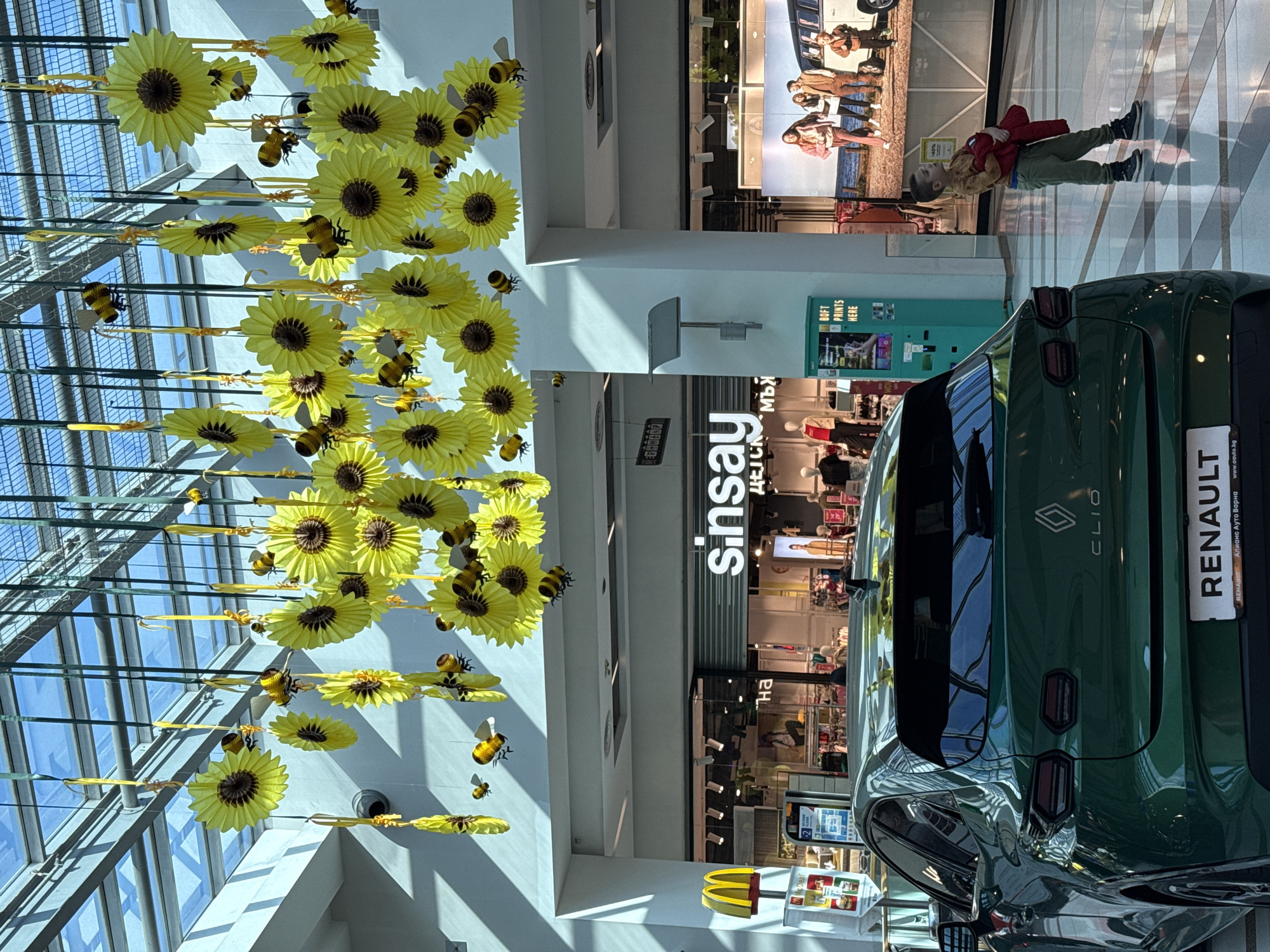 The image depicts the interior of a shopping mall, identified as being in Varna, Bulgaria, by the "Alians Auto Varna" branding on a displayed vehicle. Overhead, numerous yellow paper sunflowers and bumblebee ornaments are suspended from a glass ceiling structure, casting soft shadows. In the foreground, a dark emerald green Renault Clio hatchback is prominently displayed, seen from the rear, with its taillights, "CLIO" badging, and "RENAULT" and "Alians Auto Varna" license plate frame clearly visible.
To the left of the car, a partial McDonald's sign is visible. In the background, a "Sinsay" clothing store is clearly visible with its storefront open, displaying merchandise and mannequins. To the right of the car, a young child, appearing to be a boy, stands on a light-colored tiled floor. He wears a brown long-sleeved shirt, olive-green pants, and dark sneakers, holding a red garment or object in his left arm, and is looking upwards towards the ceiling decorations. Further to the right, a large digital display panel shows an outdoor scene with a family standing next to a classic Volkswagen van. A turquoise self-service kiosk, labeled "SOFT PRINTS HERE," stands between the displayed car and the large digital screen. The mall interior is brightly lit, likely by natural light through the glass roof.