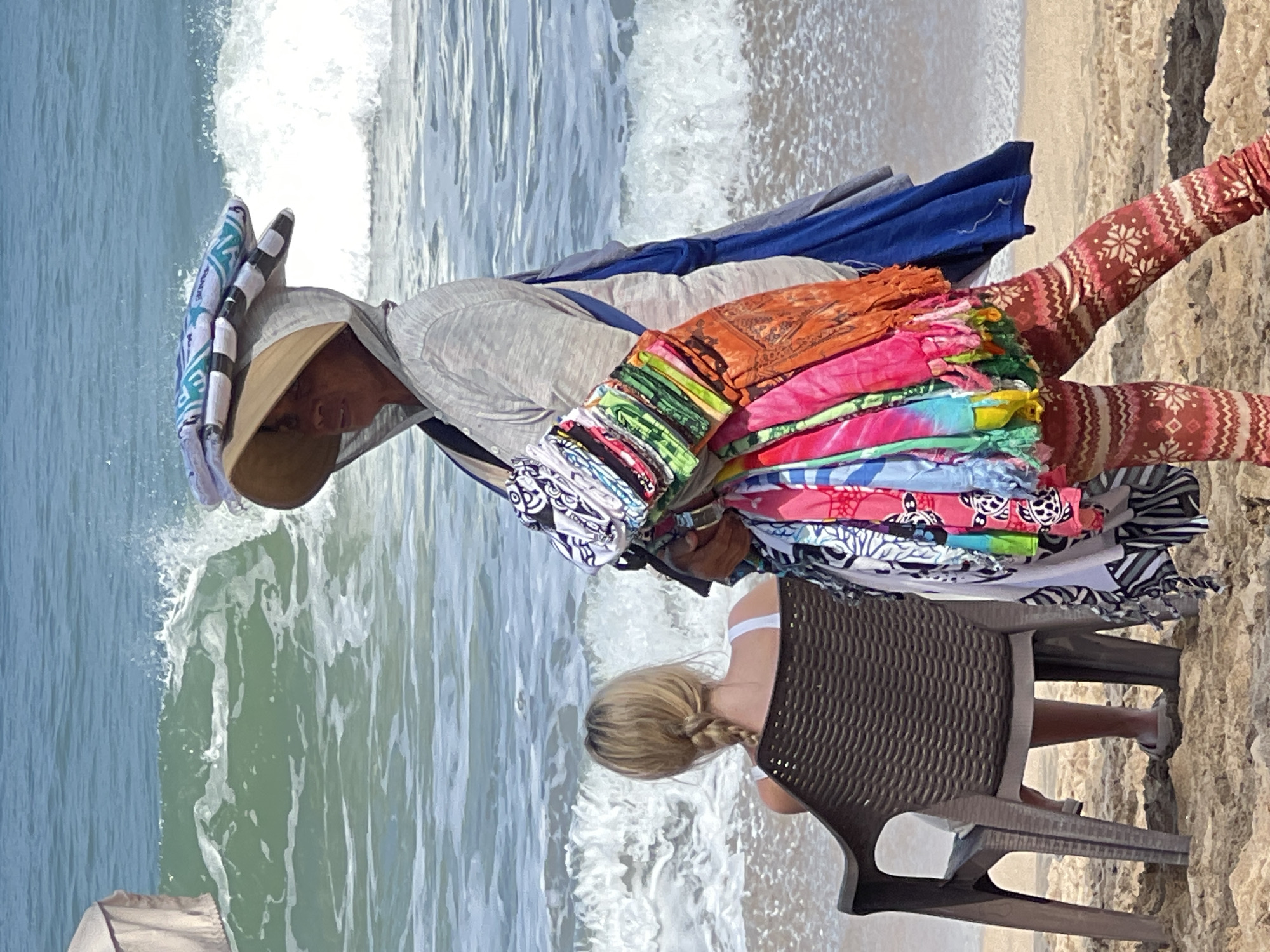 A vendor walks along the beach in Kuta, Indonesia, carrying a load of colorful textiles. She wears a wide-brimmed hat, a grey hooded jacket, and patterned leggings. In her arms, she holds a stack of vibrantly colored scarves and sarongs. To her left, a young woman with blonde hair in a braid sits in a brown plastic chair facing the ocean, wearing a white top. The background shows the turquoise water and white waves of the Indian Ocean. The sand is light brown. A partial view of a light-colored beach umbrella is visible on the far left.