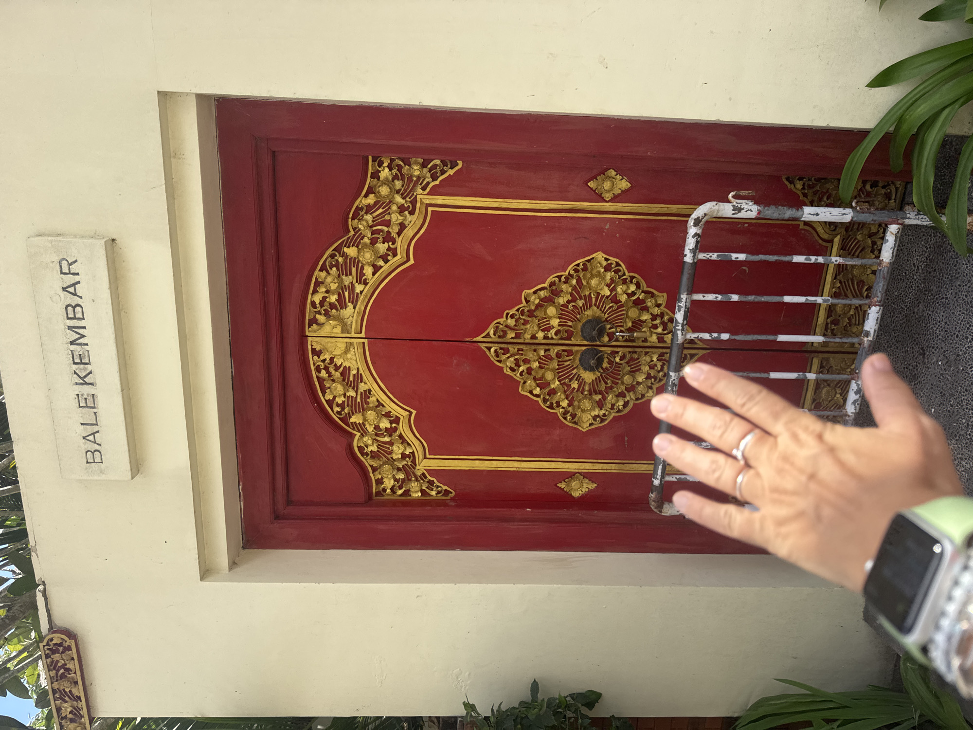In Kuta Selatan, Indonesia, a person's hand, adorned with two rings and a smartwatch, is held up in front of a closed, ornate red double door. The doors feature intricate gold detailing and are partially obscured by a metal barrier. Above the door, a sign reads "BALE KEMBAR". The wall surrounding the door is painted a light cream color, and lush green plants are visible on either side of the entrance. The scene appears to be an outdoor setting with natural lighting.