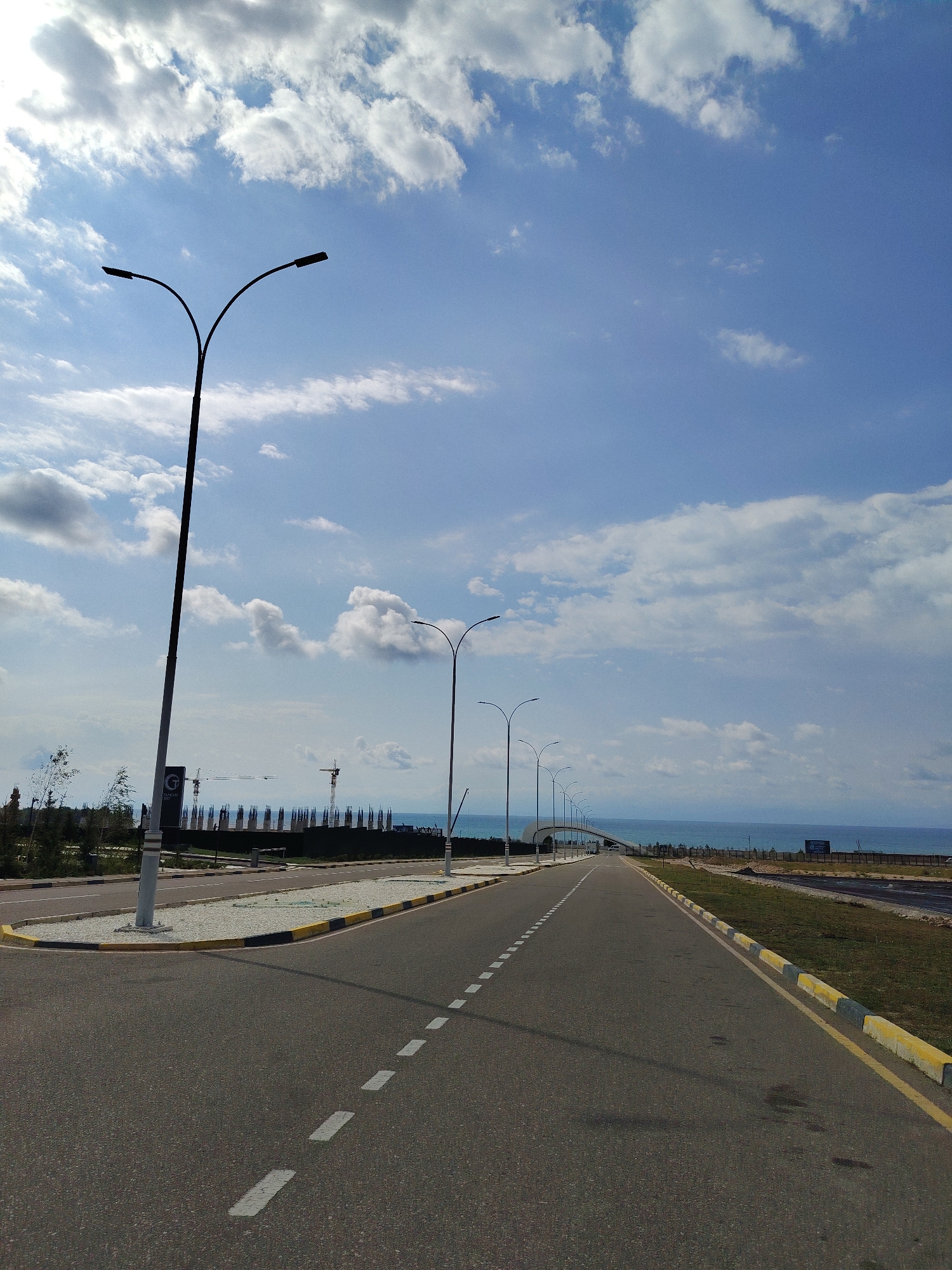 The image depicts a road in the city of Choktal, Kyrgyzstan, stretching towards the horizon. The sky is blue with scattered white clouds. Several streetlights line the right side of the road, their dark silhouettes stark against the sky. The road itself is asphalt with white dashed lines marking the lanes. On the left side of the road, there is a sidewalk and some vegetation, including trees. In the distance, construction appears to be underway, with cranes and scaffolding visible. Beyond the construction site, the blue waters of a body of water, likely Issyk-Kul Lake, are visible. The overall scene suggests a developing area or a coastal road under construction.