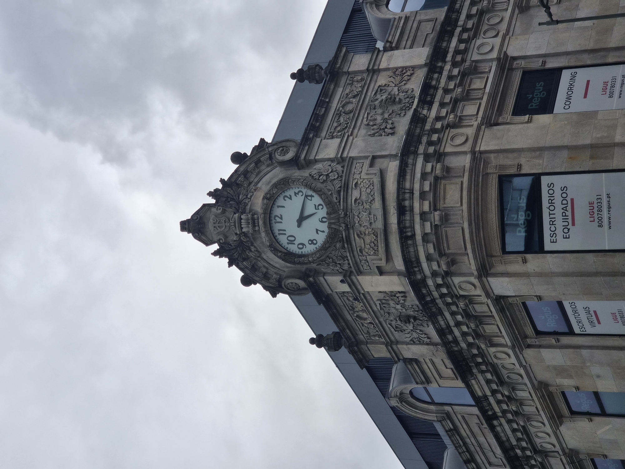 The image shows the ornate facade of a building in Braga, Portugal, featuring a large, classical clock at the top. The clock has Roman numerals and ornate hands, indicating the time is approximately 9:22. The building is constructed of light-colored stone with intricate carvings and architectural details, including decorative reliefs and moldings. Below the clock, there are windows with signs for "Regus," advertising virtual offices and coworking spaces. The sky above is overcast and cloudy. The overall impression is of an older, distinguished building in an urban setting.