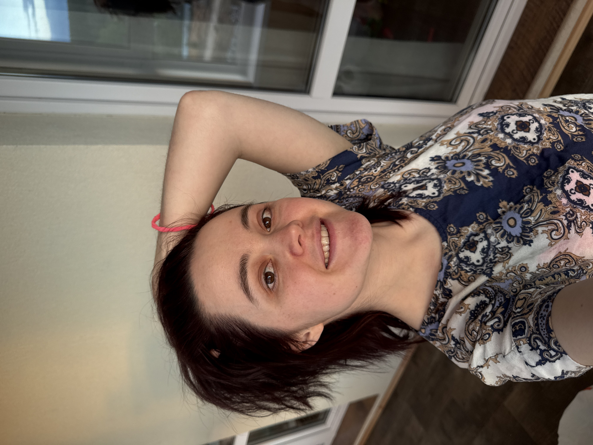 A woman with dark hair and fair skin poses indoors. Her arm is raised above her head, and she is smiling. She is wearing a patterned top. A window is visible behind her. The location cannot be determined from this image.