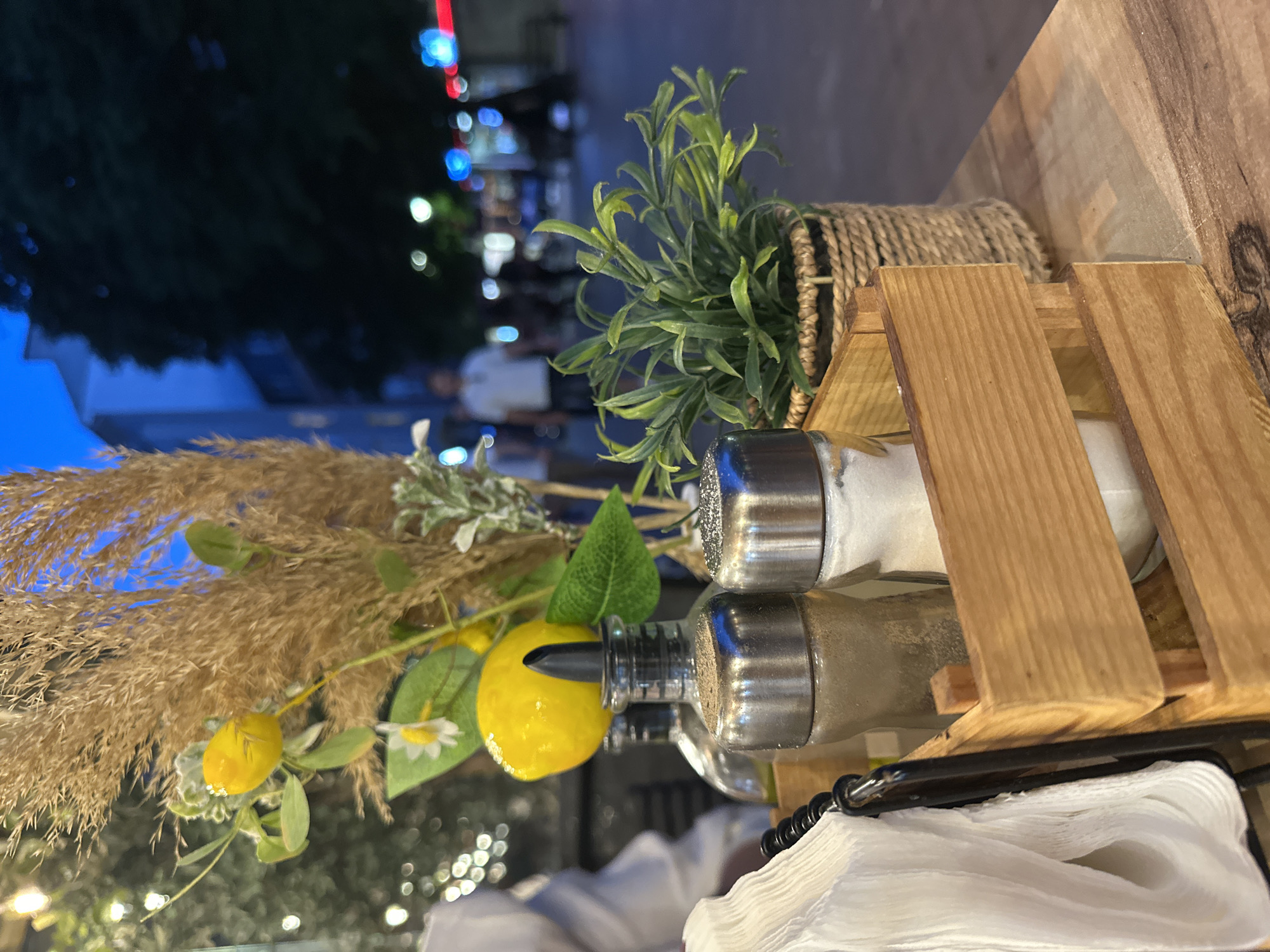 A close-up, eye-level shot captures a dining table setting at dusk in an outdoor urban location. The foreground is dominated by a wooden crate containing salt and pepper shakers, a napkin holder with white folded napkins, and two glass bottles, one with a black pour spout and the other filled with a light-colored liquid, possibly oil or vinegar. A decorative element featuring a faux lemon, small white flowers, and dried grasses rises from behind the shakers. To the right, a small potted plant with green leaves sits in a woven basket. The table's surface is made of wood. In the background, blurred figures of people are visible walking on a street lined with trees and buildings. Streetlights and illuminated signs add to the ambient lighting, creating a bokeh effect. The sky is a deep blue, indicating twilight.