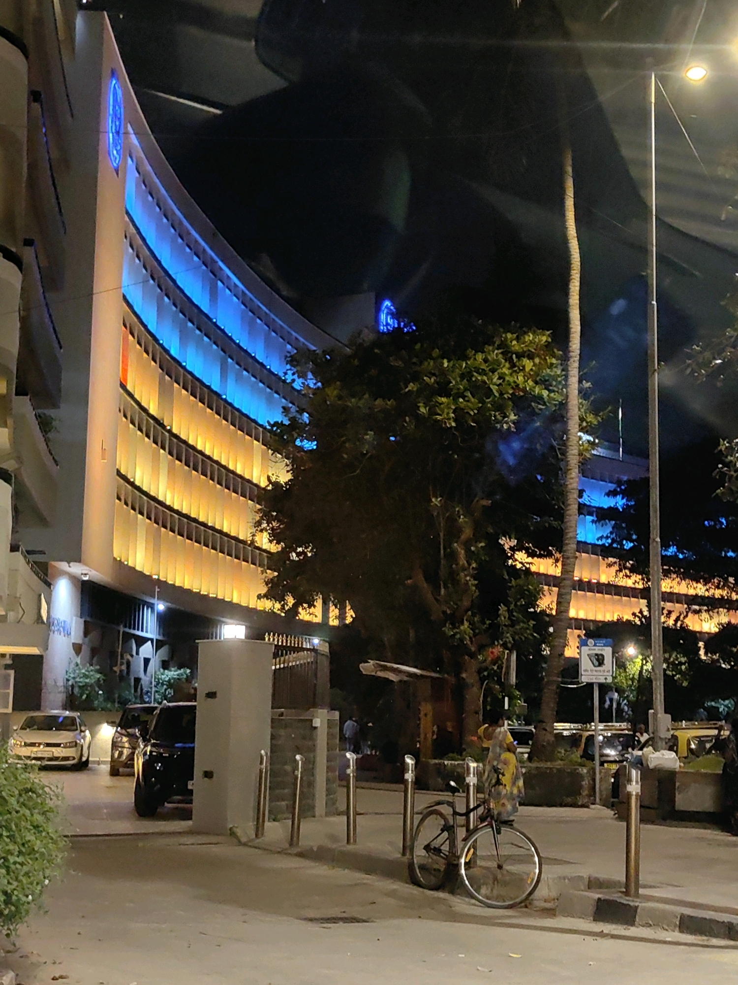 This image depicts a nighttime street scene in Mumbai, India, likely on a curve of Marine Drive, given the architectural style of the building and the characteristic lighting.
In the foreground, a bicycle with its frame and wheels visible is parked upright on a sidewalk. A person wearing a yellow and patterned garment stands near the bicycle, their back to the camera. To the left, a white car is parked facing forward. Behind it, a black SUV is parked, with its side visible.
The midground is dominated by a large, curved building with a distinctive facade. The building is illuminated with alternating bands of bright blue and orange-yellow light, creating a vibrant, modern aesthetic. The light emanates from behind vertical louvers or windows. A tall, slender palm tree stands in front of the building, its trunk dark against the illuminated facade. A street lamp with a glowing orange light is visible to the right of the palm tree.
In the background, more trees and foliage are present. Another building, also illuminated with blue and orange lights, is visible further down the street. Several other vehicles, including what appear to be taxis, are also present in the background, suggesting the area is a public thoroughfare. The overall environment suggests a well-lit urban area at night.