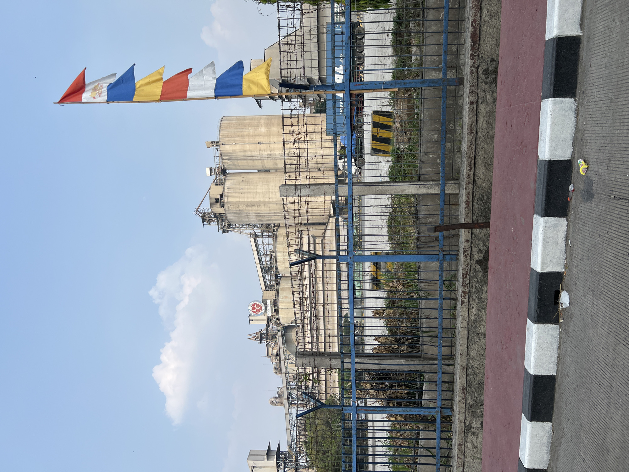This is a photograph of the PT Indocement Tunggal Prakarsa Tbk. cement factory in Citeureup, Indonesia. In the foreground, there is a road with a red and white striped curb, and several pieces of trash on the road. A blue metal fence separates the road from the factory grounds, which are covered in dry grass and weeds. Behind the fence, a large industrial complex with several silos and processing structures is visible. A flagpole with several colorful flags is also present in front of the factory. In the background, a truck with a blue container is parked in the factory yard. The sky is clear with a few white clouds.