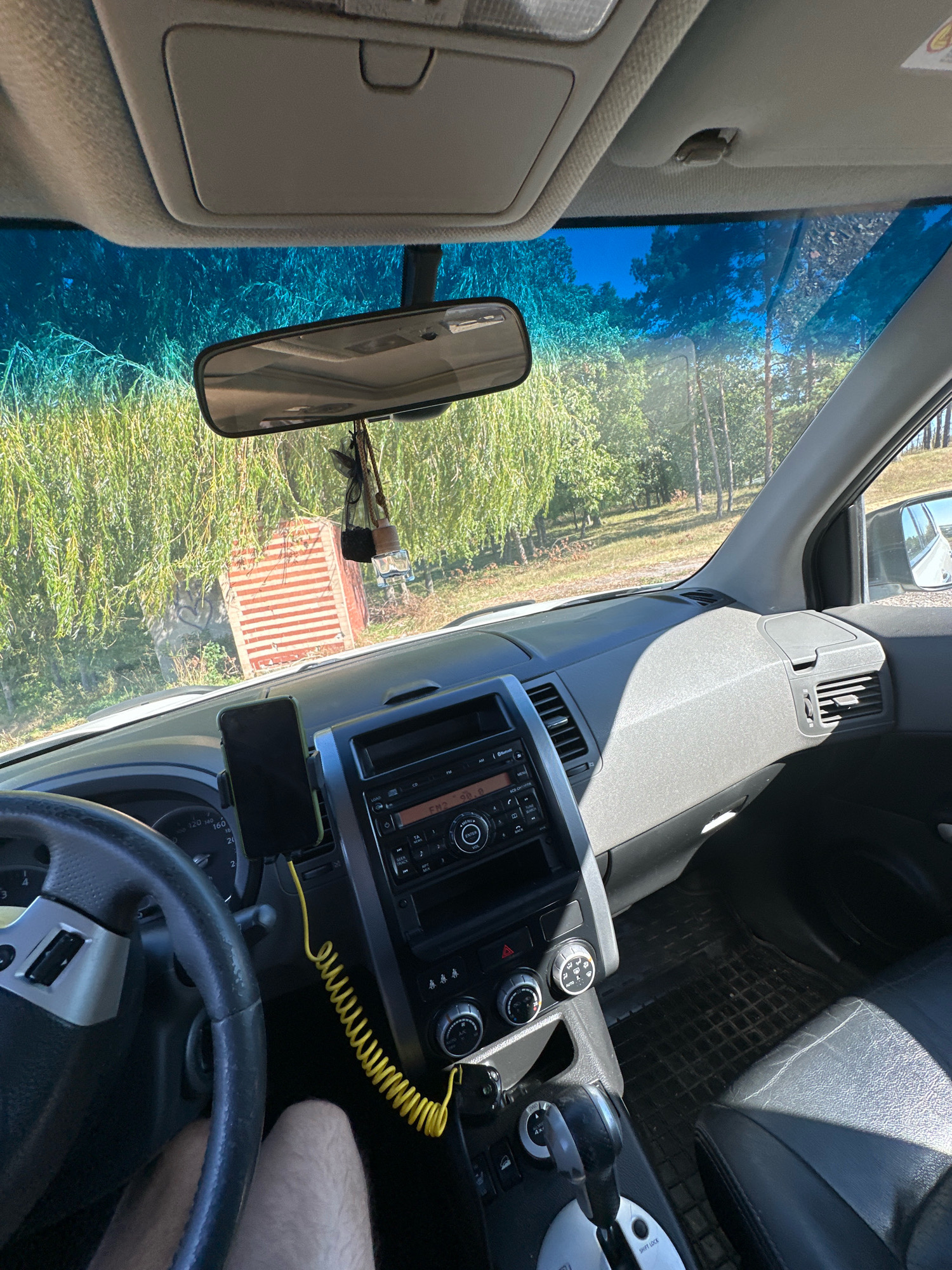 The image shows the interior of a car, looking out through the windshield and side windows. The dashboard, steering wheel, and center console are visible. A smartphone is mounted on the dashboard. Outside the car, trees and foliage are seen, along with a building with red and white stripes. The sky is blue. The image was taken in Makiv, Ukraine.