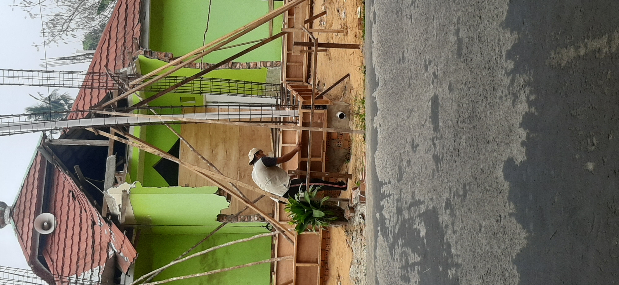 The image depicts a construction site, likely in Jambi, Indonesia, where a building is undergoing renovation or reconstruction. A person, wearing a light-colored vest over a dark shirt and a light-colored cap, is actively working on the foundation, which is enclosed by wooden formwork. The building itself is painted a vibrant green and has a damaged red tile roof. Structural reinforcements, consisting of metal rebar and wooden scaffolding, are visible around the building. In the foreground, a wet asphalt road reflects the overcast sky, suggesting recent rain. A small green plant is situated near the construction area. The overall scene indicates a disruptive but ongoing construction process in what appears to be an urban or semi-urban setting.