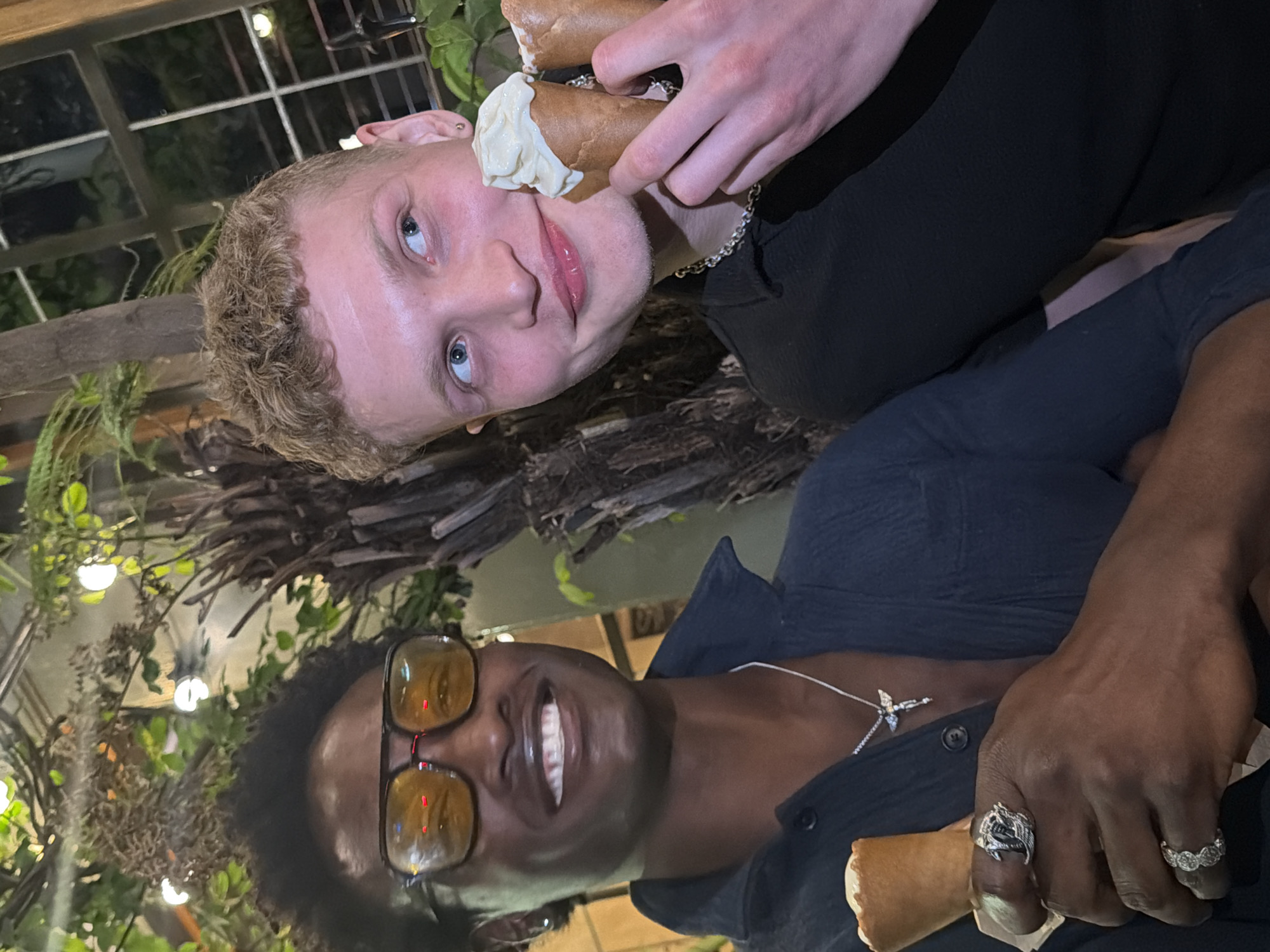 The original image is rotated 90 degrees clockwise. When viewed correctly, it depicts two individuals in a relaxed, dimly lit setting, likely an indoor or covered outdoor establishment such as a restaurant or cafe, consistent with an evening or nighttime scene in London, United Kingdom.
In the foreground, a dark-skinned individual, appearing male, is seen from the chest up, smiling broadly towards the camera. They have dark curly hair and are wearing large yellow-tinted sunglasses, a dark open-collared shirt with a silver cross necklace, and multiple rings on their fingers, including a prominent ornate silver one and two banded rings. They are holding a cannoli or similar pastry.
Behind and slightly above them, a light-skinned individual, also appearing male, is shown with a playful expression and a slight smirk directed at the camera. They have short, curly blonde hair and a beard, striking blue eyes, and pink lips. This person is wearing a dark long-sleeved top and a silver chain necklace. They are holding two cannoli, with some cream from one playfully smudged on their cheek/nose area.
The background features lush green foliage and intertwining wooden or branch-like structures, creating a natural, garden-like aesthetic. Above, a structure with square panes, possibly a skylight or roof, with visible artificial lights, suggests an enclosed space. Both individuals appear happy and are engaged in a lighthearted moment, enjoying their pastries in a convivial atmosphere. There is no visible text in the image.