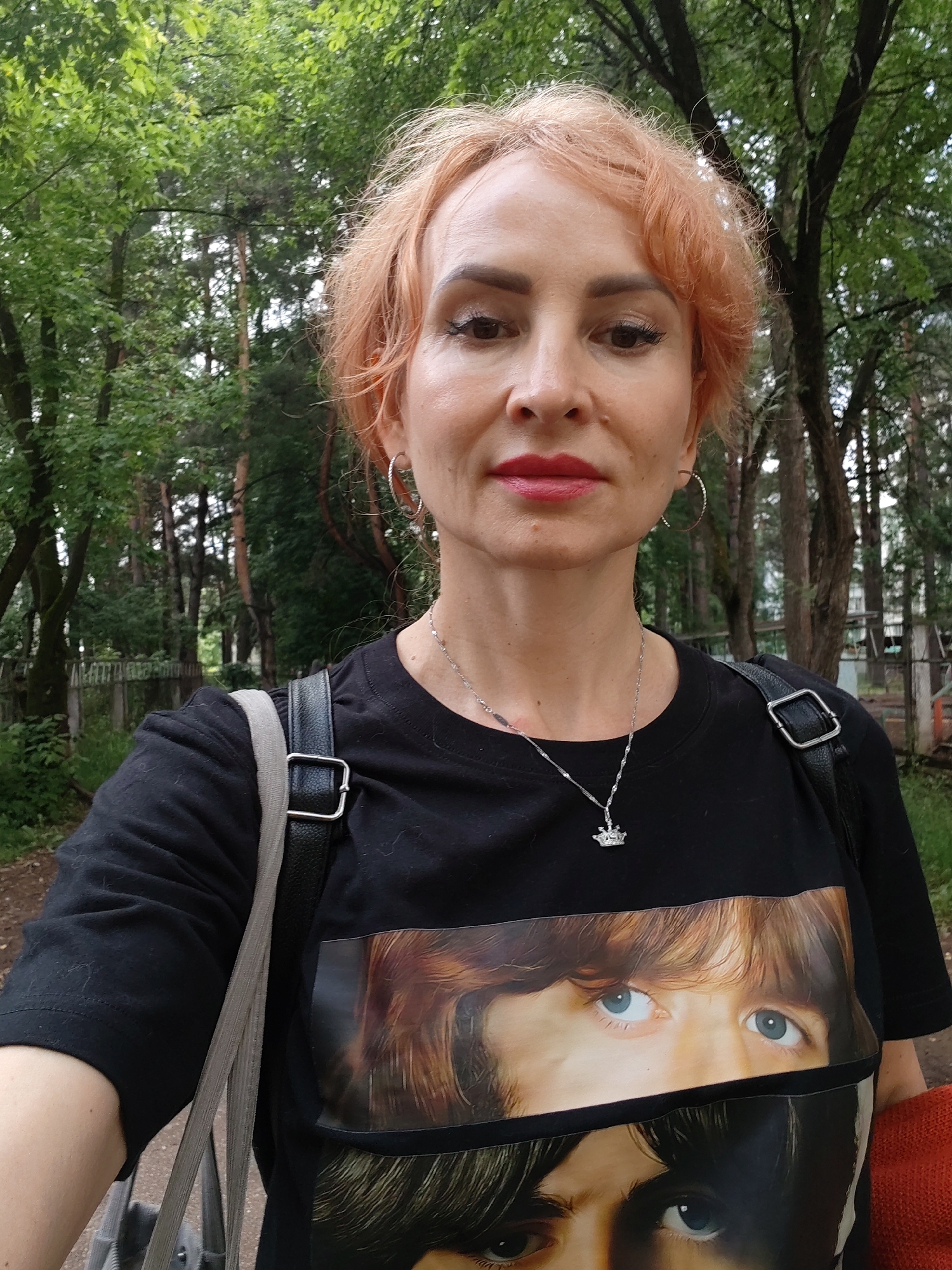 A middle-aged Caucasian woman with short, peachy-pink hair and fair skin is captured in a medium-shot selfie. She is wearing a black t-shirt with a graphic print of a close-up of eyes and hair, possibly from a band or celebrity. A silver necklace with a crown pendant rests on her collarbone. She has on hoop earrings and subtle makeup, including red lipstick and eyeliner. Her expression is neutral, and she appears to be outdoors in a park-like setting with trees and foliage in the background. To the left, a bag strap is visible. The environment suggests a temperate climate. The background features a wooden fence and more trees, indicating a natural or semi-natural area. The image might have been taken in Chaikovsky, Russia, given the context.