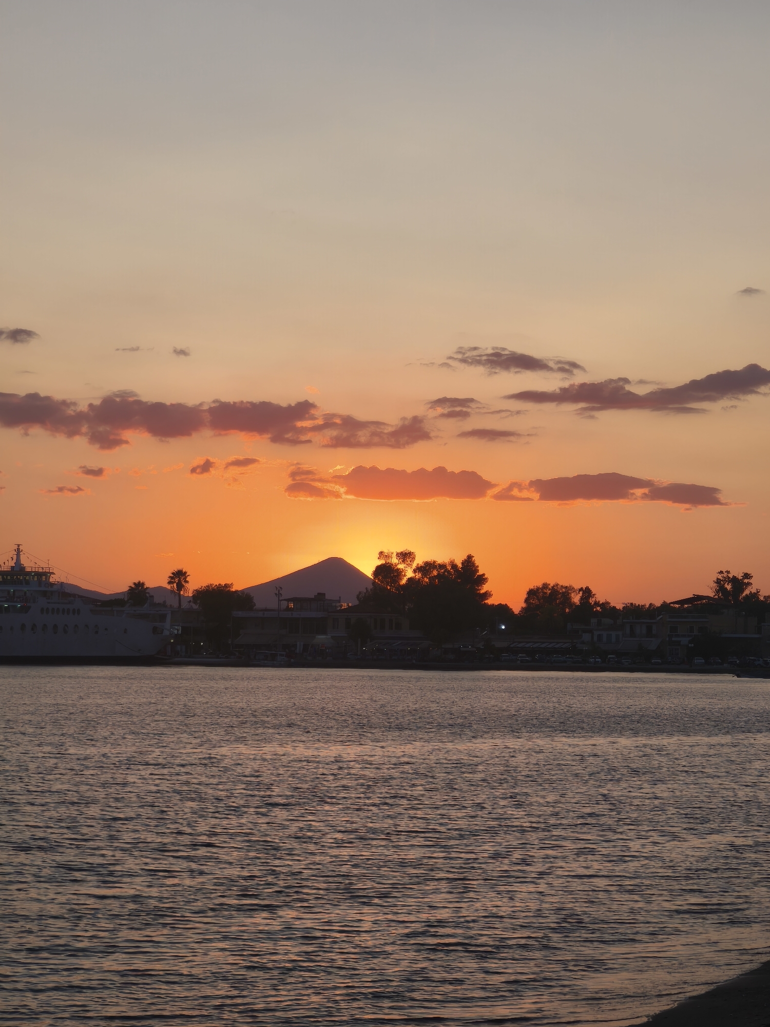 A coastal scene at sunset in Eretria, Greece, features a large ferry docked on the left, a body of water in the foreground, and a silhouetted town and mountain range in the background. The sky displays a gradient of colors from light grey at the top to vibrant orange and pink near the horizon, with scattered clouds. The water is choppy and reflects the warm hues of the sky. Buildings and trees are visible along the shoreline, and the outlines of a mountain are discernible behind the town. No people are clearly visible.