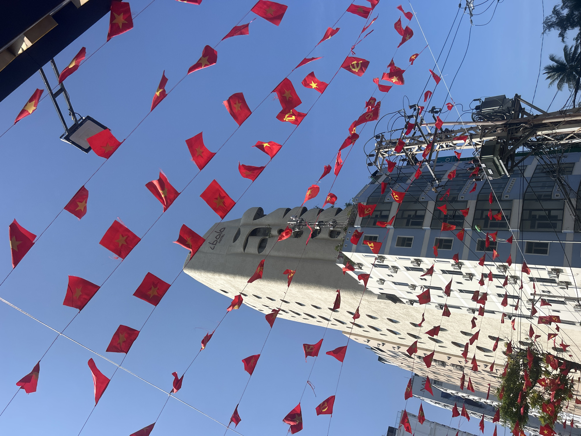 The image shows a street scene in Bắc Nha Trang, Vietnam. The view is directed upwards towards the sky, partially obscured by numerous small red flags hanging on strings. These flags appear to be Vietnamese national flags, each featuring a yellow star. Some flags are slightly tattered.
In the background, two buildings are visible. One is a modern structure with rounded edges and numerous recessed oval openings, seemingly a hotel or commercial building, which bears the inscription "Lbala Hotel". The other building, taller and partially obstructed by the flags, has a more conventional design with rows of windows.
Power lines and electrical equipment are also visible between the flags and buildings, suggesting an urban setting. The sky is clear and blue. No people are visible in the frame.
