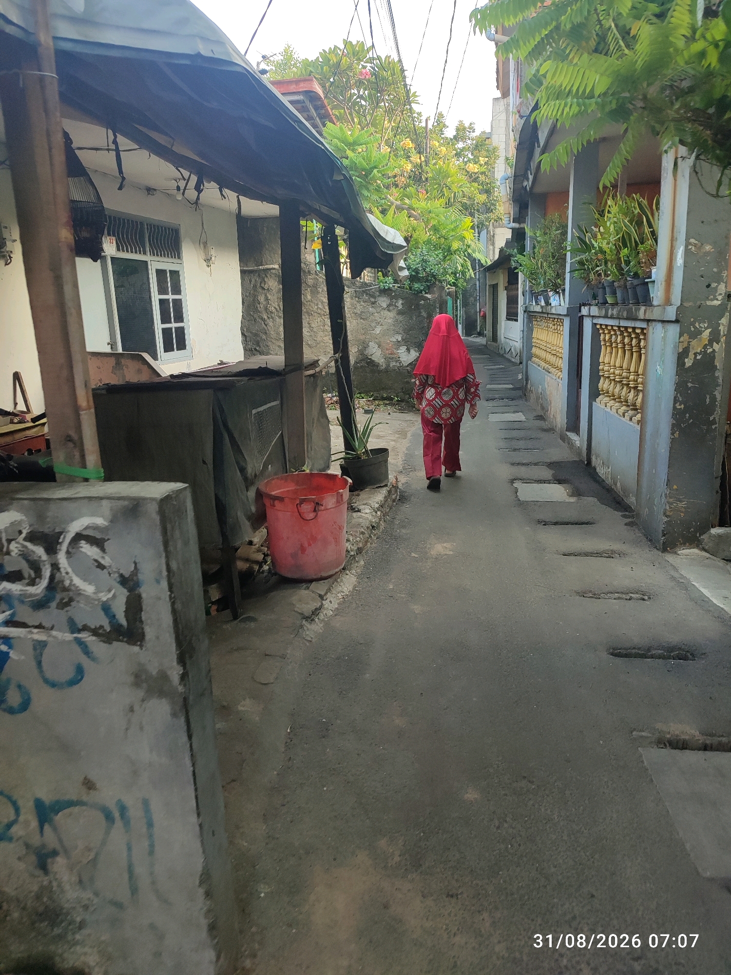 The image depicts a narrow alleyway in East Jakarta, Indonesia, bathed in the soft light of early morning, as indicated by the timestamp "31/08/2026 07:07." A person wearing a vibrant red hijab and matching patterned attire walks away from the camera down the paved alley. The alley is flanked by buildings with simple facades, some showing signs of wear and tear. To the left, a white building with a window is partially visible, shaded by an improvised awning. A birdcage hangs from the awning. A large red bucket stands near a covered structure. On the right side of the alley, a building features a decorative yellow railing and potted plants. The ground of the alley is asphalt with several rectangular indentations, possibly drainage covers. Graffiti is visible on a concrete structure in the foreground on the left. Lush green foliage from trees and plants is abundant, softening the urban environment. The overall impression is of a residential area in a developing city, with a mix of simple structures and natural elements.