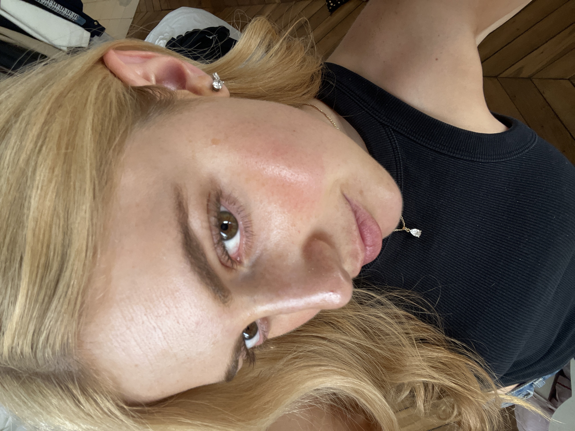 A blonde-haired woman is seen from a close, angled perspective. She has fair skin, light brown eyes, and is wearing a black top. She is also wearing a diamond earring and a diamond necklace. The background includes glimpses of a wooden floor, some clothing items, and neutral-toned bedding. The woman's face is slightly flushed. There is no information that suggests the location of the picture is in Paris, France.