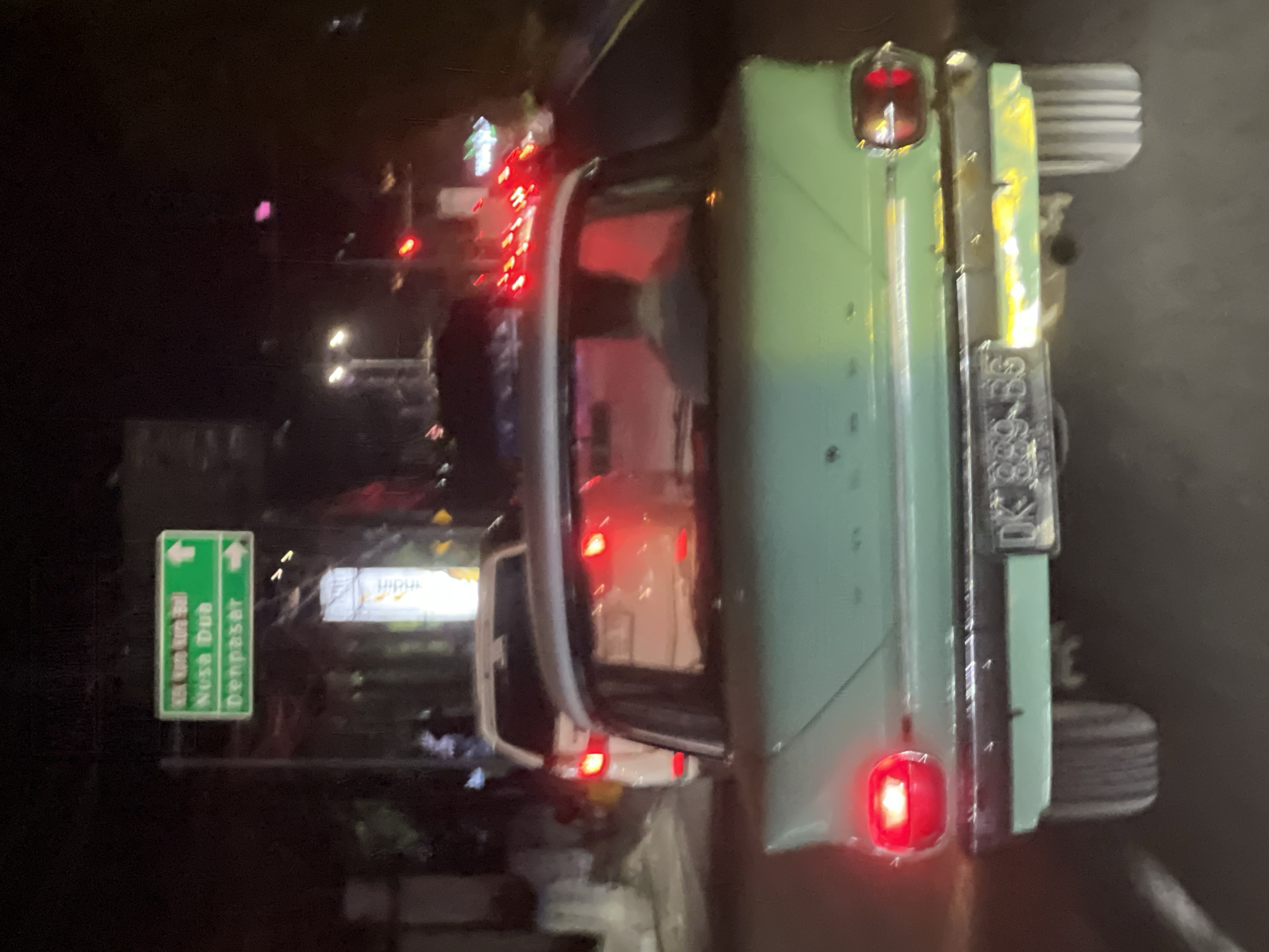 A green vintage car, illuminated by its red taillights, is on the road. Its license plate reads "DK 889 BG." A green road sign points towards Nusa Dua and Denpasar. The setting appears to be the city of Denpasar, Indonesia, at night. Other cars are visible, lit with red lights. A street light can be seen in the background, along with other light sources in the distance.