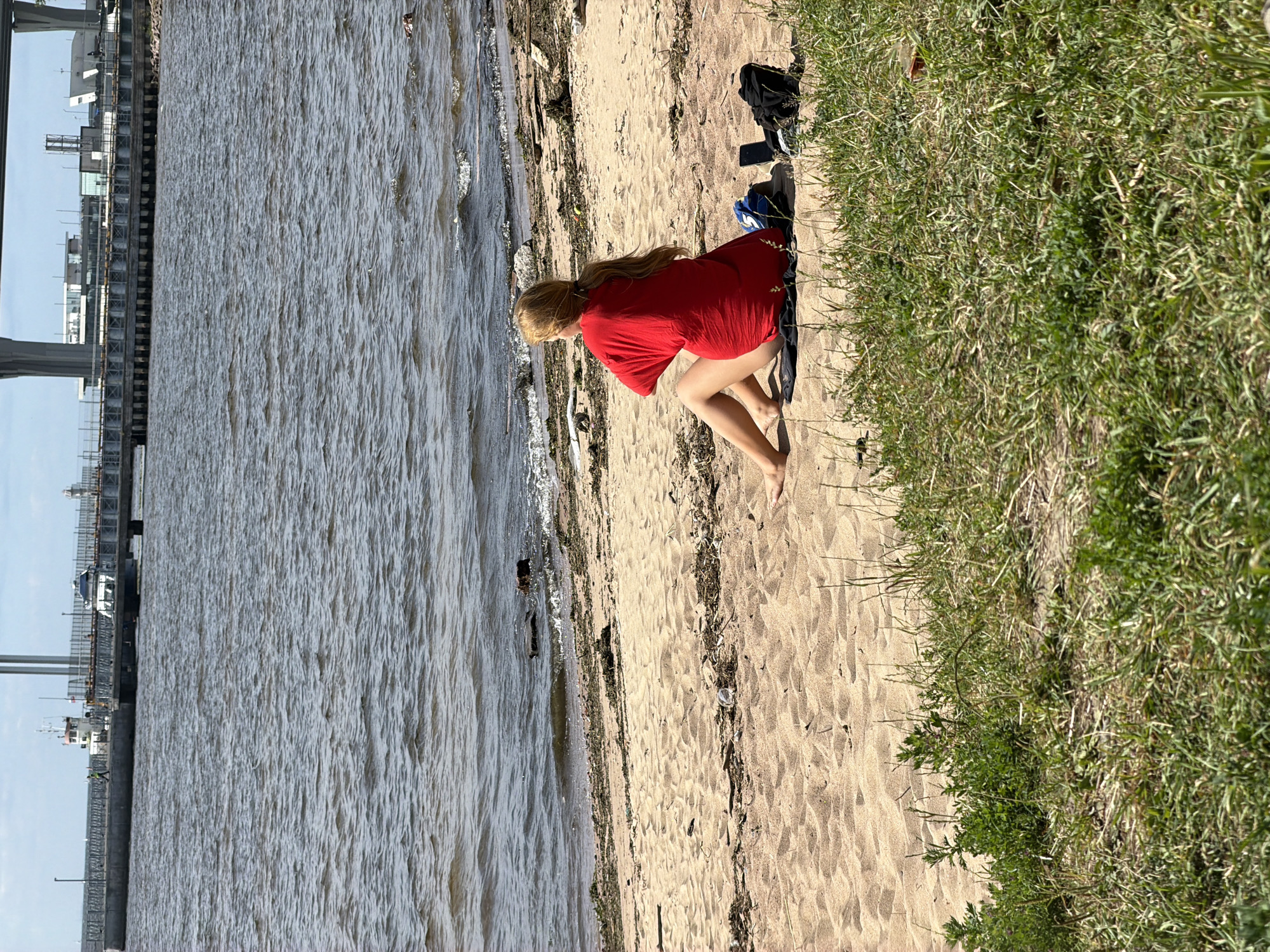 A young woman with blonde hair pulled back in a ponytail is sitting on a sandy beach, facing away from the camera. She is wearing a bright red, oversized t-shirt and appears to be bare-legged. Her legs are crossed, with her right foot resting on the sand. Next to her on the sand are a dark blue bag, a black backpack, and a smartphone. The beach is lined with sparse green vegetation in the foreground. In the background, the choppy, grey water of a bay stretches out, with a pier or bridge structure visible on the horizon. Several vessels, including what appears to be a tugboat and another ship, are docked or moving near the structure. The sky is overcast. The scene is likely on the Gulf of Finland or one of its connected waterways within or near Saint Petersburg, Russia, given the presence of the large industrial and infrastructural elements in the background, common to the city's waterfront areas.