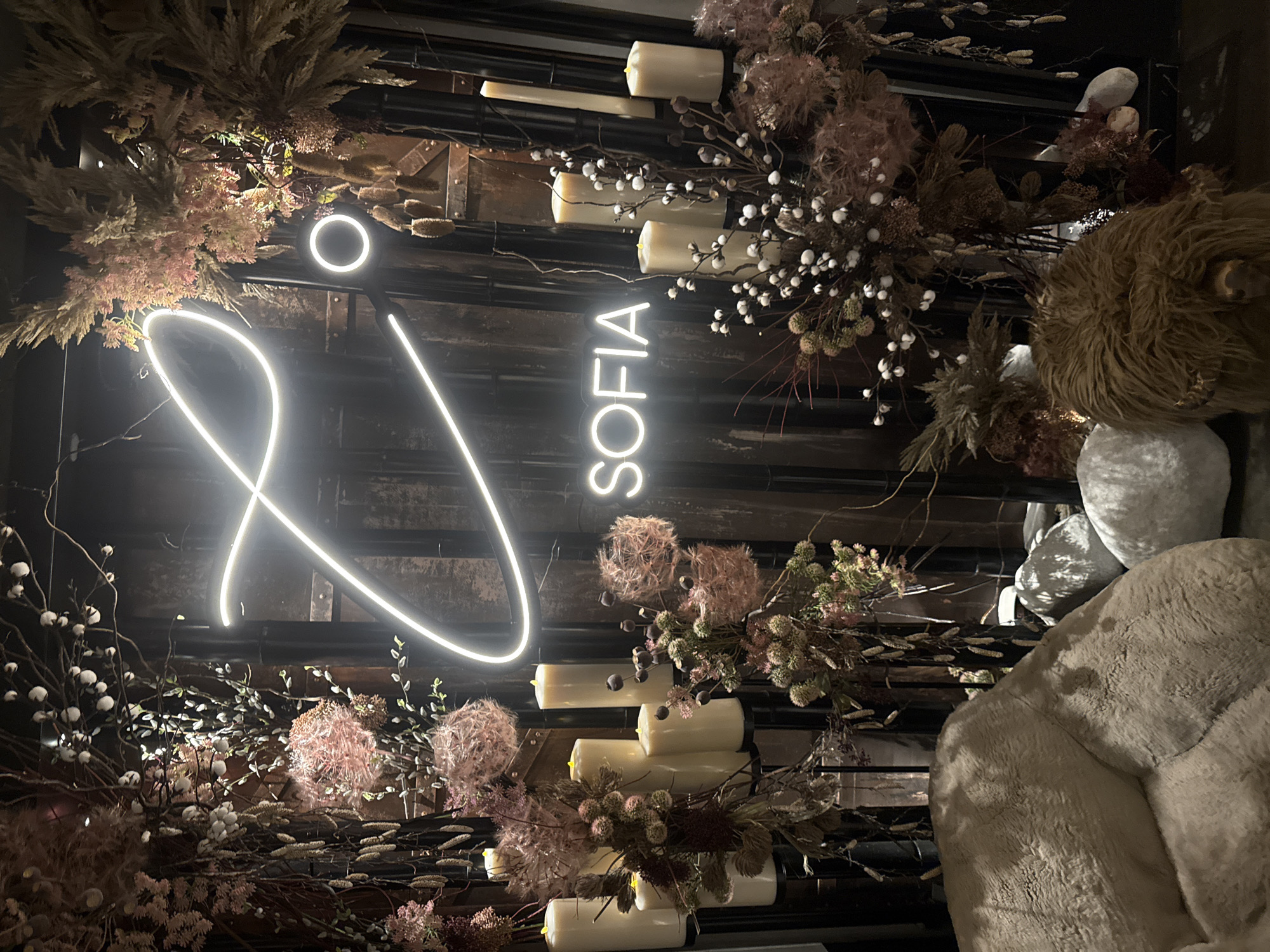 The image depicts a dimly lit interior decorated with dried floral arrangements, an illuminated neon sign spelling "SOFIA," and several pillar candles. In the foreground, a plush, cream-colored beanbag chair and a brown, shaggy decorative object resembling a highland cow are visible, alongside smooth, grey stones. The background features a dark wooden wall with metal accents, behind which the neon sign is mounted. The overall ambiance suggests a trendy establishment, possibly a bar or restaurant, located in Sofia, Bulgaria, given the prominent signage. No individuals are present in the frame.