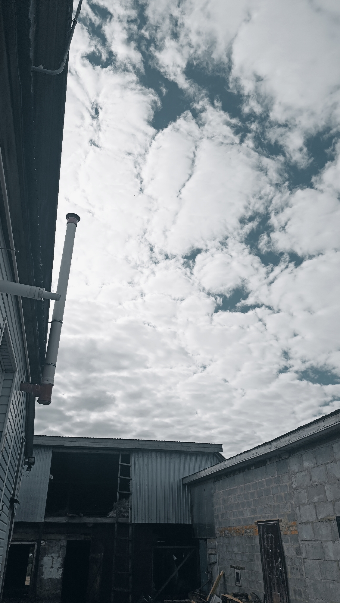 The image shows an outdoor scene with a cloudy sky. In the foreground, to the left, a section of a building with vertical siding and a visible pipe system, including a tall, light-colored pipe with red accents, is present. The main structure in the middle ground is a large building with corrugated metal siding, featuring an open, dark interior with visible internal structures like ladders and debris. To the right, another building constructed from gray concrete blocks is partially visible, with a dark doorway. The overall scene appears to be industrial or utilitarian, possibly a construction site or an abandoned facility. The location within the city of Karsha, Russia, is not visually indicated by any specific landmarks or signage in the image.