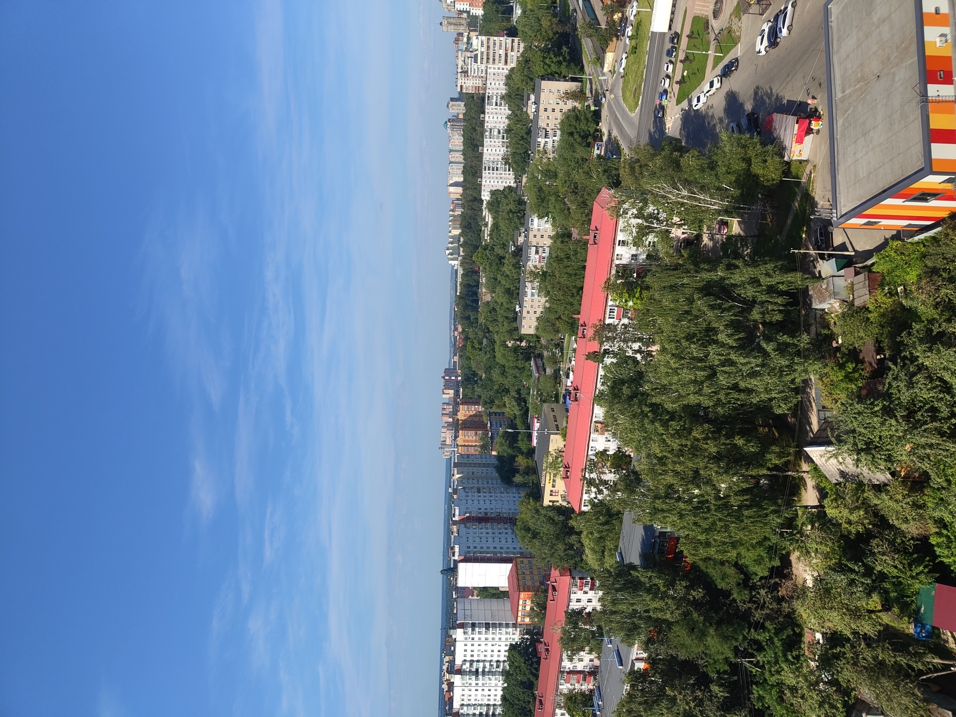 The image captures a high-angle view of a residential area in Ramenskoye, Russia, on a clear, sunny day with a bright blue sky and scattered wispy clouds. The urban landscape is characterized by a mix of multi-story apartment buildings and smaller residential structures, interspersed with lush green trees and vegetation.
Prominently featured are several apartment buildings with predominantly white and light-colored facades, some with red accents and roofs. One larger building in the foreground on the left has a white facade with numerous windows, and adjacent to it are buildings with red roofs. Another building with a red roof is situated centrally in the mid-ground. Further in the distance, a collection of taller apartment buildings, in various shades of white, beige, and orange, forms the skyline.
A winding road with parked cars is visible in the right side of the image, suggesting moderate urban activity. The presence of trees, some quite tall and mature, indicates a well-treed environment within the city. Smaller, older-looking residential buildings and garages are nestled amongst the trees in the lower left portion of the frame. A colorful, modern-looking building with orange, yellow, and white stripes is partially visible in the bottom right corner. The overall scene depicts a typical day in a Russian residential district.