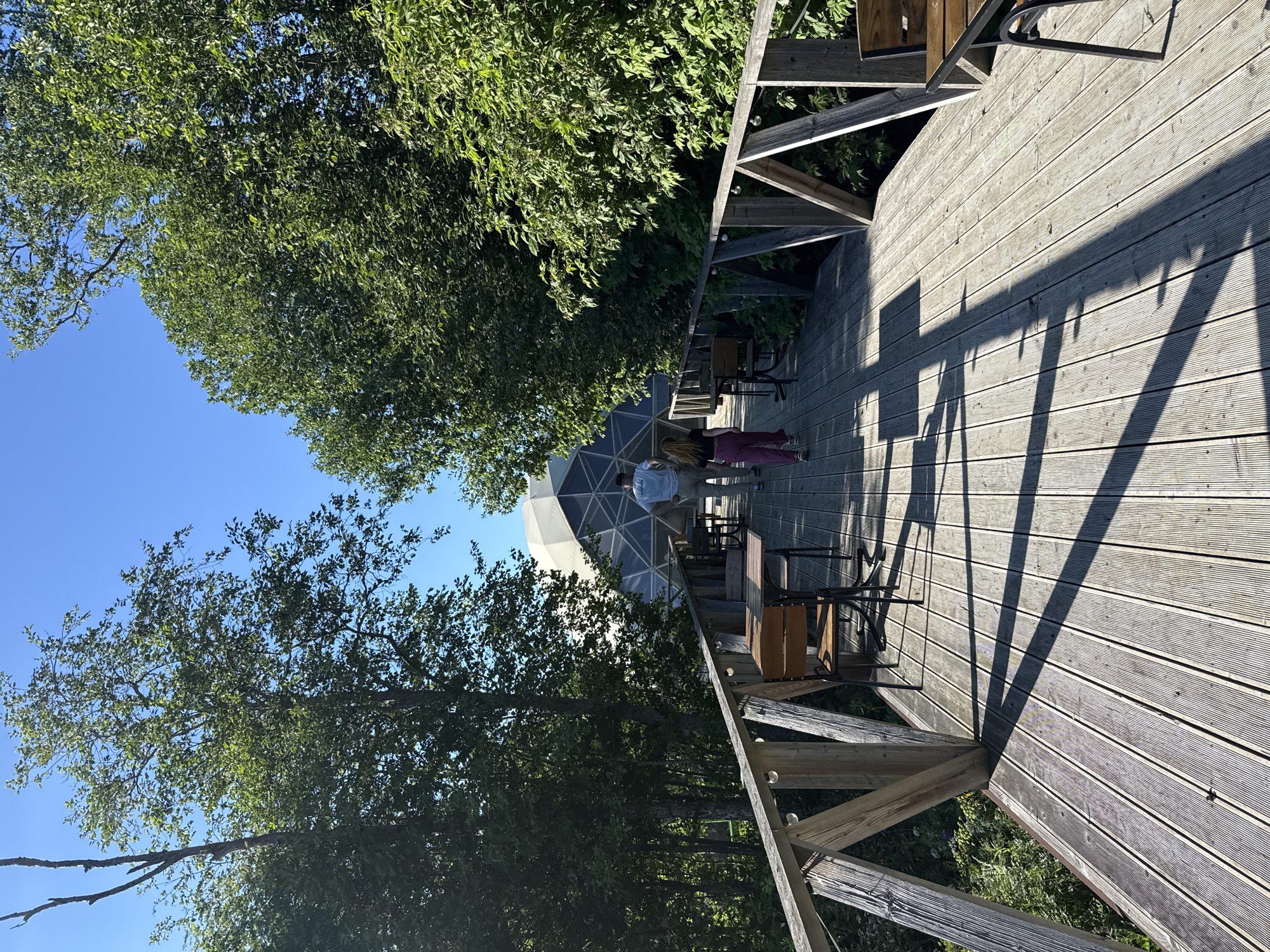 A wooden boardwalk with tables and chairs leads towards a geodesic dome structure. Two individuals, a man and a woman, are walking away from the camera on the boardwalk. The scene is surrounded by lush green trees under a clear blue sky. The location is identified as the city of Zamosh'e (Zaklinskoe S/P), Russia.