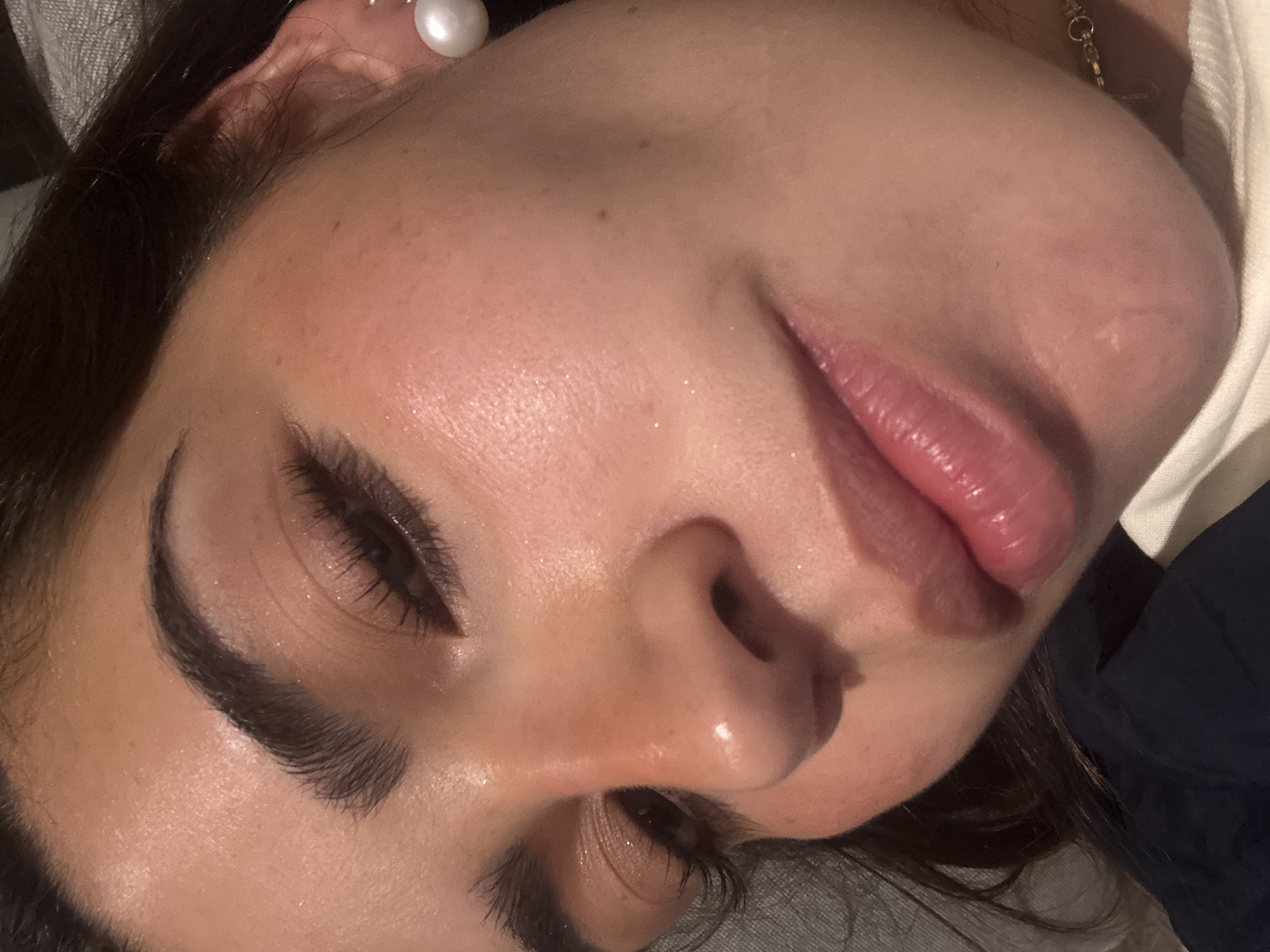 A close-up, high-angle shot depicts a person's face. The person has dark hair and fair skin, with prominent eyebrows and eyelashes. The eyes are closed. The lips are full and glossy. A pearl earring is visible in the ear. The skin appears to have a slightly oily sheen. No specific landmarks or environmental elements are discernible to indicate the location of Mexico City. The image focuses solely on the person's face and lacks contextual details.
