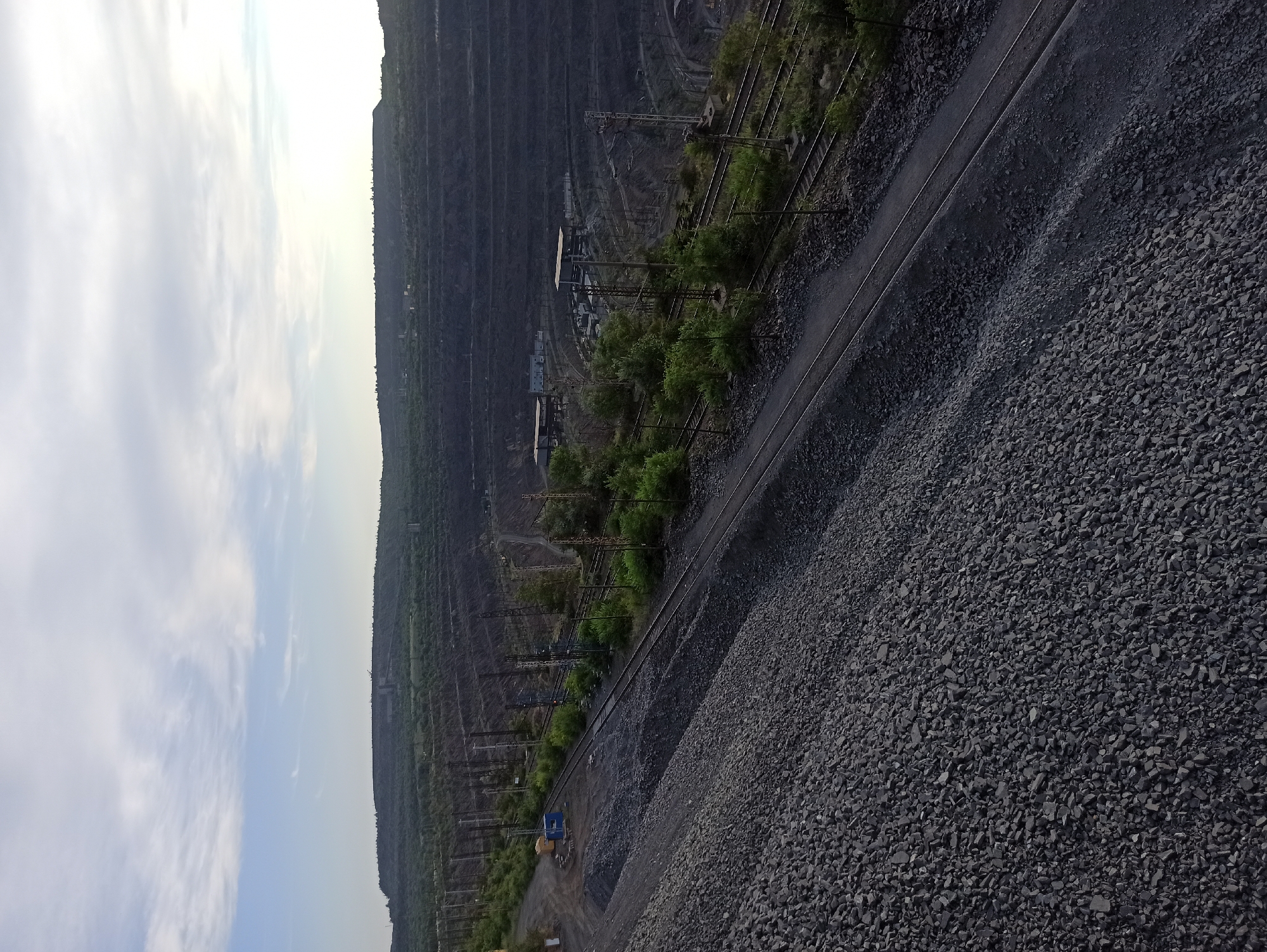 This is a vertically oriented, wide-angle photograph of a quarry or open-pit mine. The foreground is dominated by a large pile of dark grey, crushed rock, likely stone or coal. To the left of this pile, a set of railway tracks runs parallel to it, disappearing into the distance. Beyond the tracks, the landscape drops dramatically into the quarry, revealing multiple terraced levels carved into the earth.
Various structures are visible within the quarry, including buildings with metal roofs, what appear to be conveyor belts, and industrial machinery. Overhead power lines and their supporting structures crisscross the scene, indicating active industrial operations. Patches of green trees and vegetation are scattered throughout the quarry and along its edges, suggesting areas where the land is reclaiming or where vegetation is maintained.
In the far distance, the quarry walls recede, appearing as layered striations of dark earth. The sky on the far left is bright and filled with wispy white clouds against a light blue background. The sun appears to be low on the horizon, casting a soft light, suggesting it is either early morning or late afternoon. The overall mood is industrial and vast, highlighting the scale of the mining operation and the natural landscape. There is no visible text in the image.