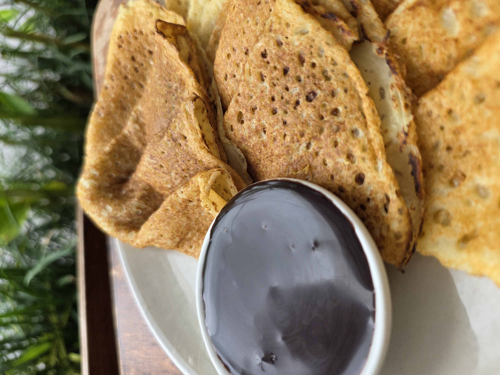 A close-up, eye-level shot shows a plate of golden-brown crepes served with a small bowl of dark, glossy chocolate sauce. The crepes are folded and stacked, with visible texture and small perforations. The chocolate sauce fills the white bowl and reflects the light. In the background, out of focus, are lush green foliage and a wooden surface. The scene appears to be outdoors, possibly on a table or tray. There are no people present in the image.