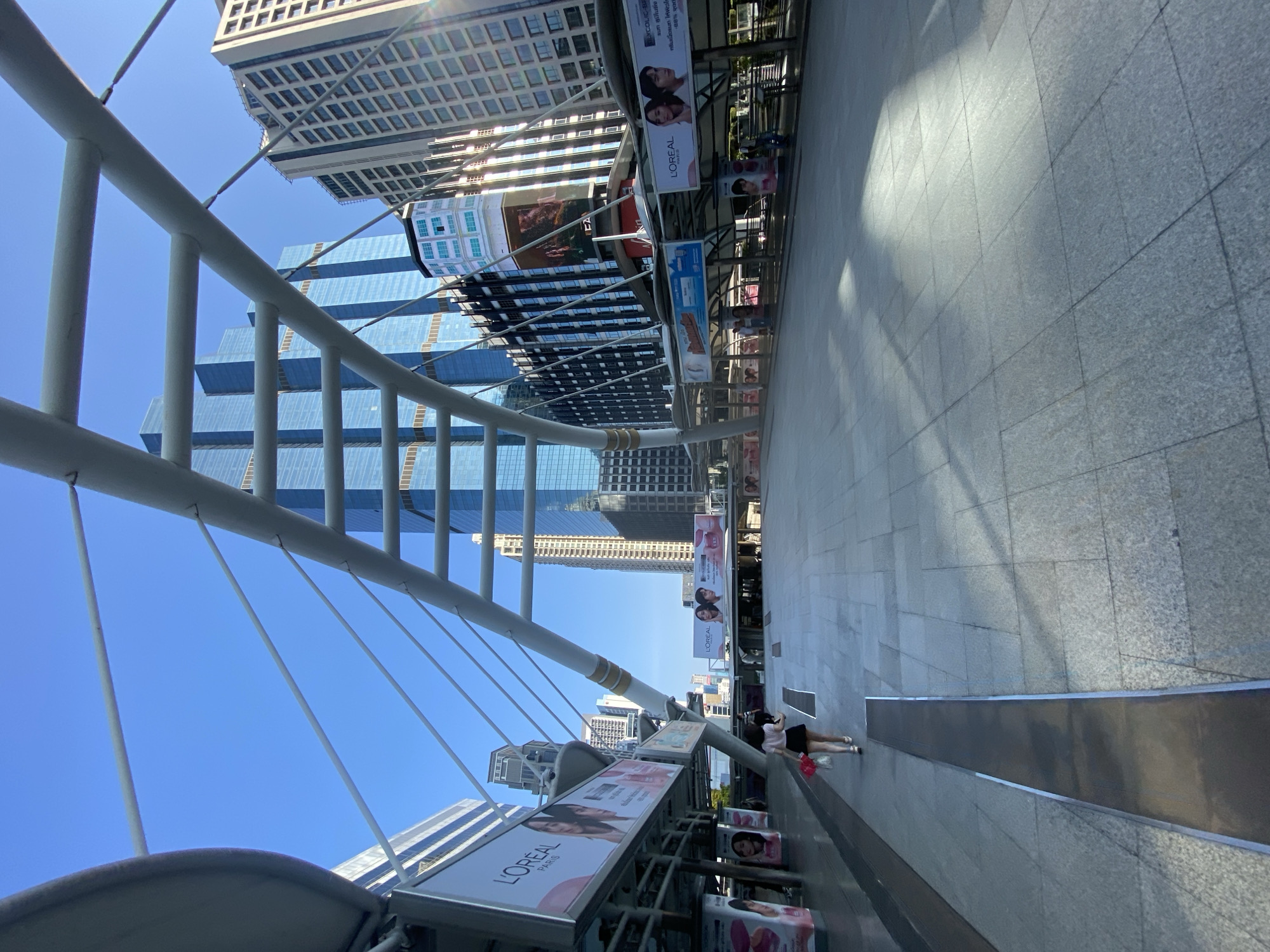 The image displays an elevated pedestrian walkway in a modern city, under a clear, bright blue sky, suggesting a sunny day, likely mid-morning or afternoon given the shadows. The perspective is looking along the length of the walkway.
In the foreground, a prominent white pedestrian bridge structure with diagonal cables extends into the distance, framing the upper left portion of the image. Tall, contemporary skyscrapers dominate the background, some featuring reflective blue glass facades, while others are constructed from lighter-toned concrete.
The walkway itself is wide and paved with grey tiles. Along the right side, multiple advertising billboards are mounted, primarily featuring beauty products. Two people, appearing to be women, are visible on the walkway in the mid-distance; one is wearing a dark skirt and light top and carries a red bag. A long, low, reflective metal barrier or bench runs along the right edge of the walkway.
Visible text on the billboards includes "L'OREAL PARIS" and "L'OREAL" repeatedly, often alongside images of women's faces. Another advertisement includes the text "GLYCOLIC BRIGHT" and Thai script, specifically "ลูมิในที" (Lumineuse), "คลีมเนื้อจนท," and "ให้หัวใ," likely detailing product information or benefits. The scene captures a typical bustling urban environment with modern architecture.
