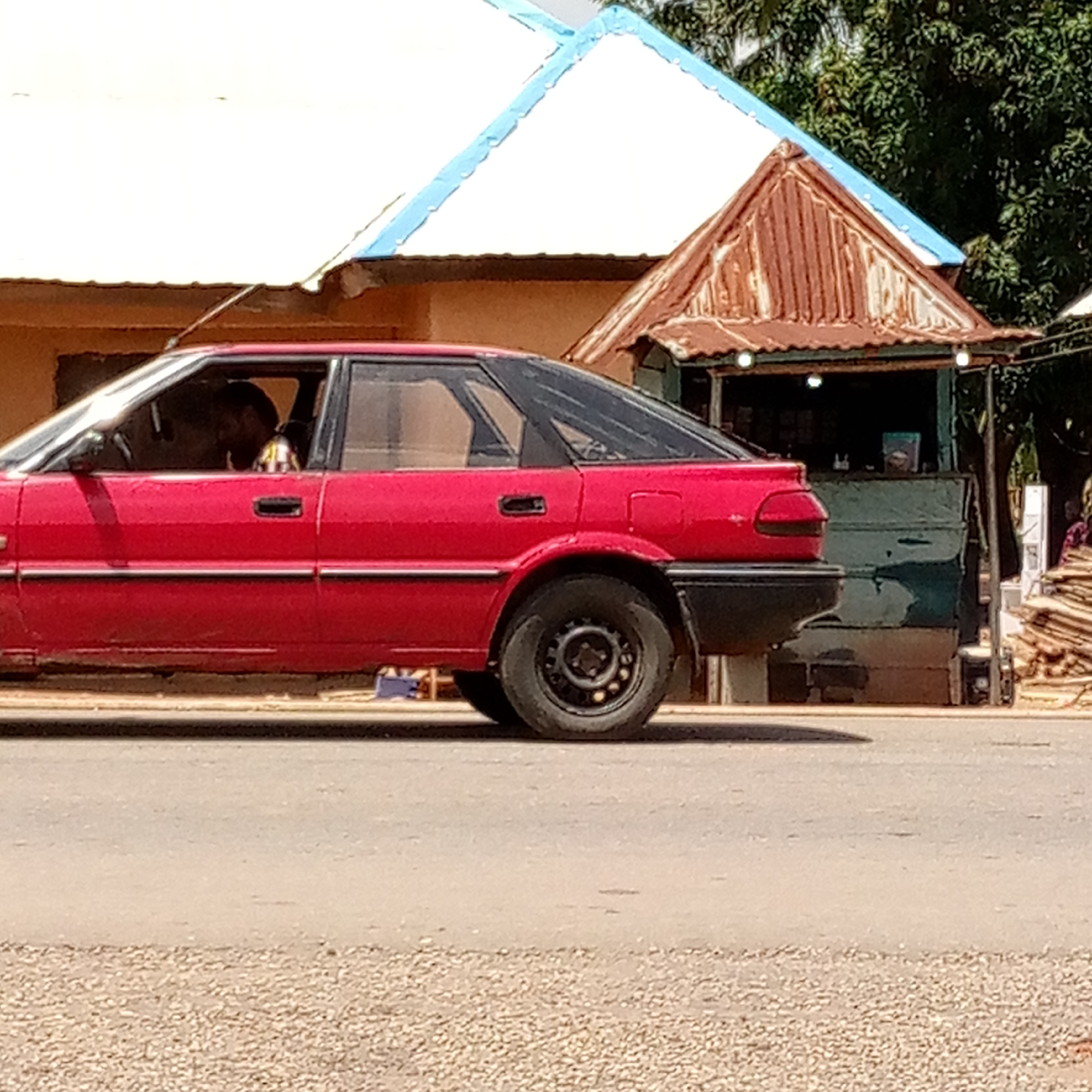 A red sedan is traveling on a road in Mararaba, Nigeria. The vehicle has black trim and black wheels. A driver is visible inside the car. Behind the vehicle are two buildings: a beige structure with a white roof trimmed in blue, and a green and corrugated metal stand. A pile of wood and foliage are also visible behind the stand. The road is paved and has a shoulder of gravel.