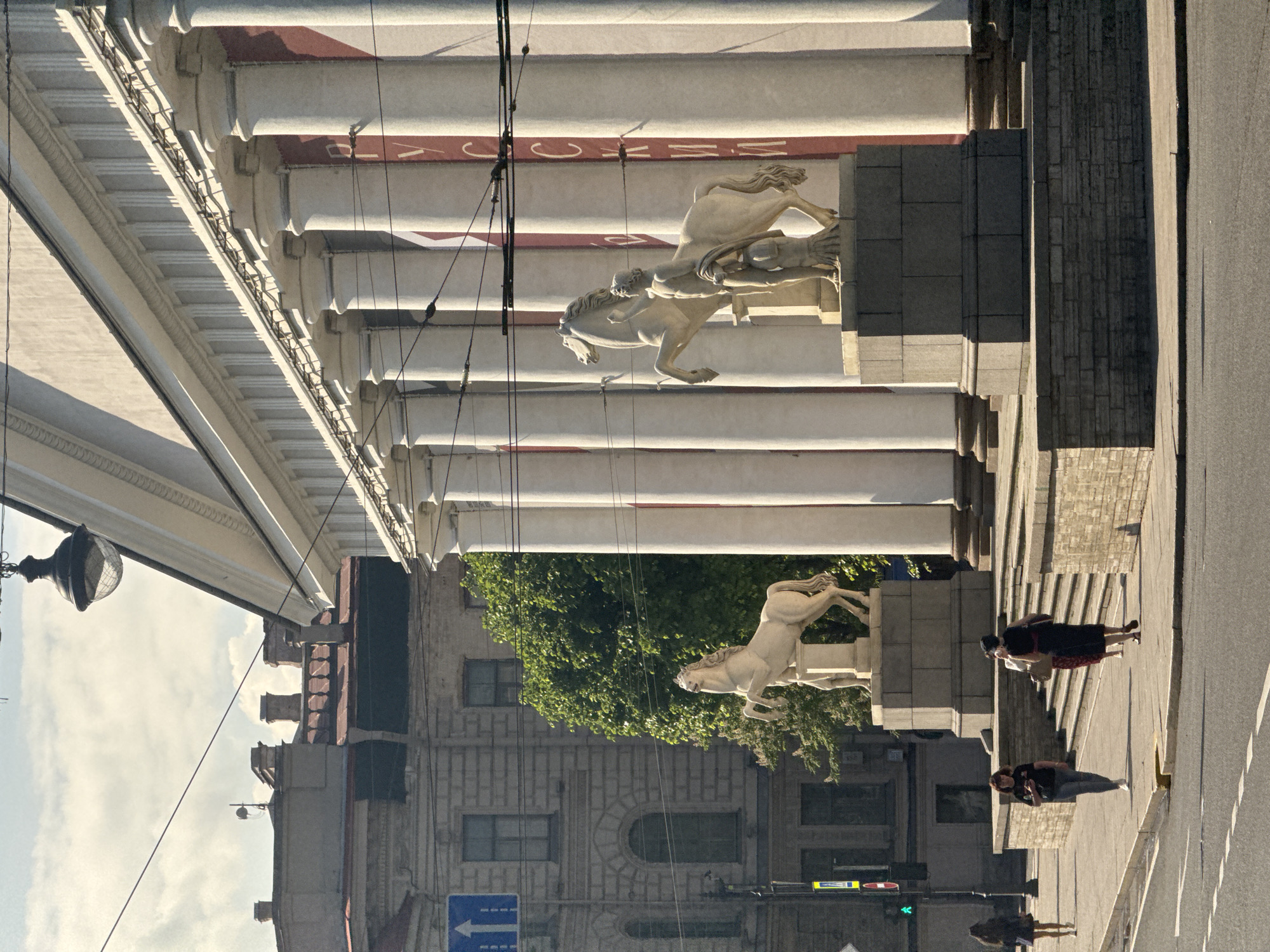 This image shows a street view of the State Russian Museum in Saint Petersburg, Russia. The museum's imposing Neoclassical facade, characterized by a series of tall white columns, dominates the right side of the frame. Banners with Cyrillic lettering are visible behind the columns. In front of the museum, two equestrian statues stand on pedestals. The left statue depicts a horse rearing on its hind legs, while the right statue shows a figure holding the reins of a standing horse, with another smaller figure at its feet.
A sidewalk runs along the base of the museum, and several people are walking on it. On the left side of the image, a multi-story building with a light-colored facade and arched windows is visible. A traffic light with a green signal and a street sign pointing upwards are also present. The street in the foreground is paved and appears to be a road with markings. The overall scene is bathed in sunlight, casting shadows on the pavement and architectural elements.