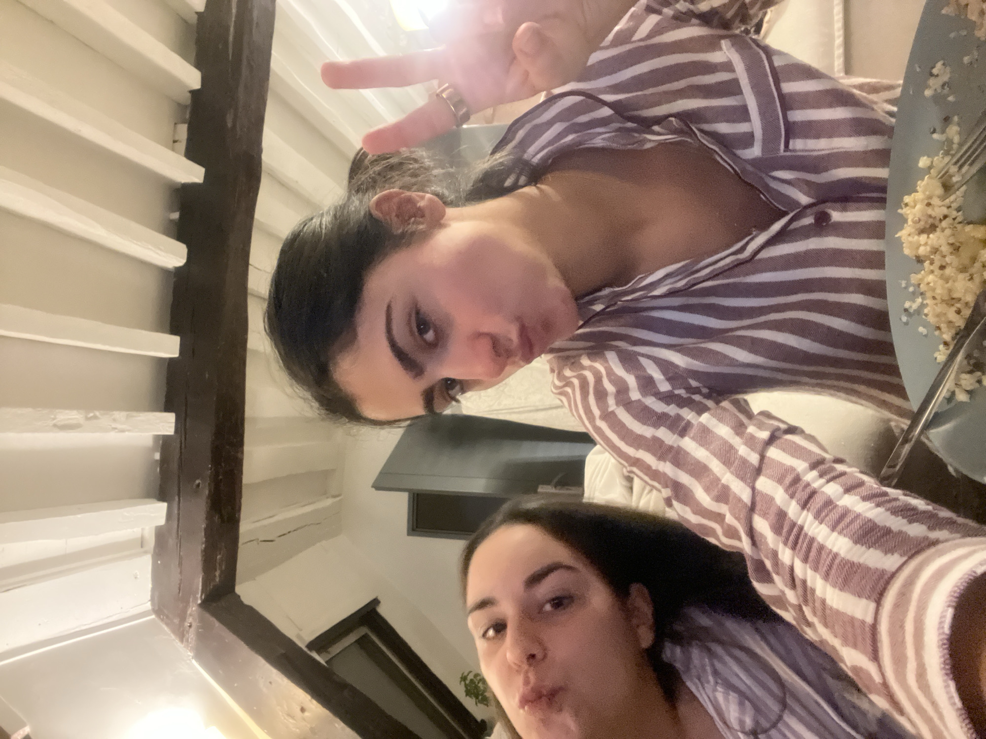 Two women pose for a selfie. One woman is in the upper portion of the frame and is holding up her fingers in a peace sign. She has dark hair pulled back, and she is wearing a striped pajama top. A plate of food and a fork are also visible. The second woman is in the lower portion of the frame. She has dark hair and is also wearing a striped pajama top. A wooden beam is visible in the background. The location appears to be indoors, possibly an apartment in Paris, France.