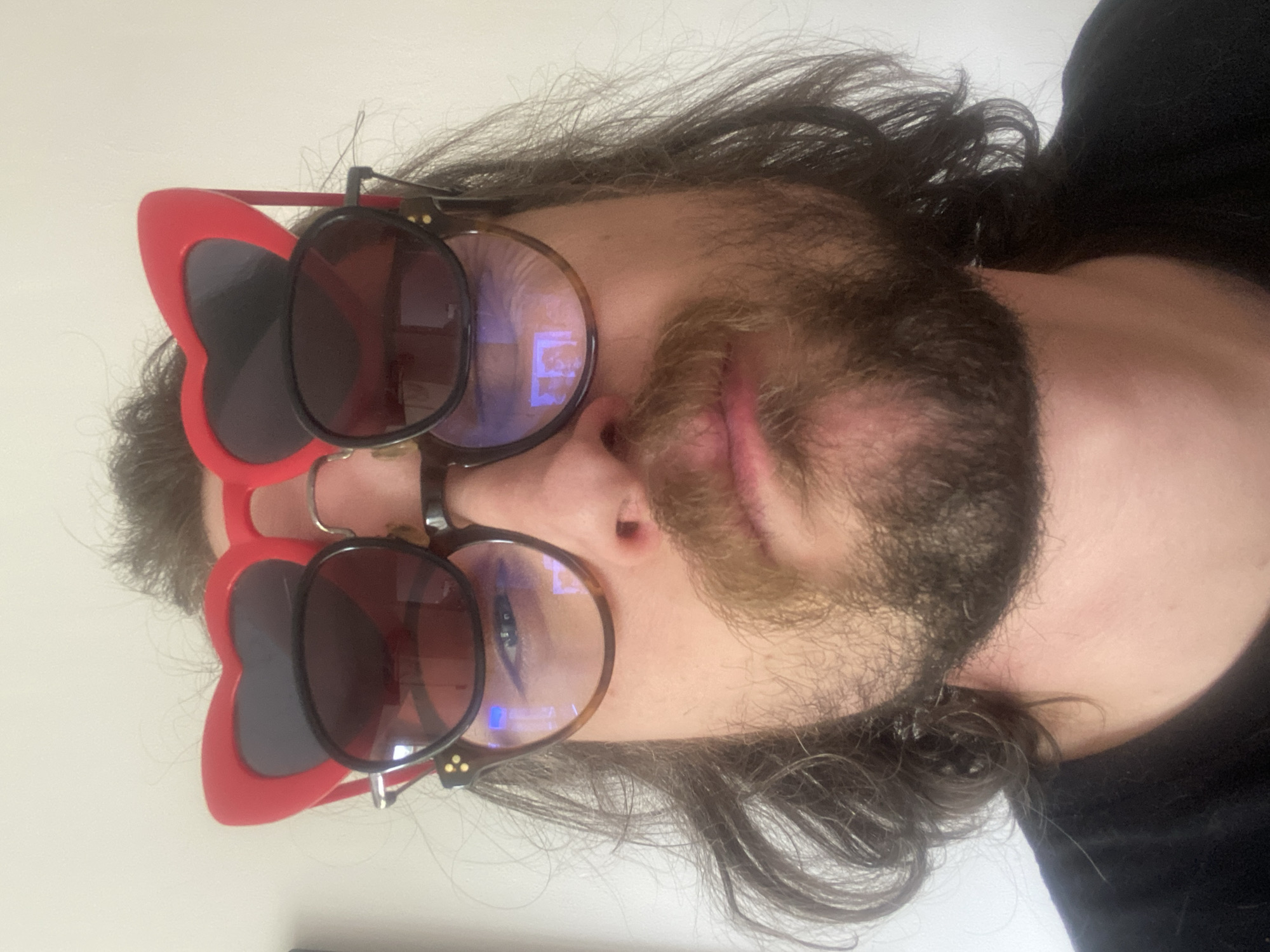 The image shows a person with long brown hair and a beard. They are wearing eyeglasses with clip-on sunglasses attached. The sunglasses are red and heart-shaped. The background is a plain white wall. The image is taken from a low angle, looking up at the person. There is no indication of the location being Rusanovo, Russia.