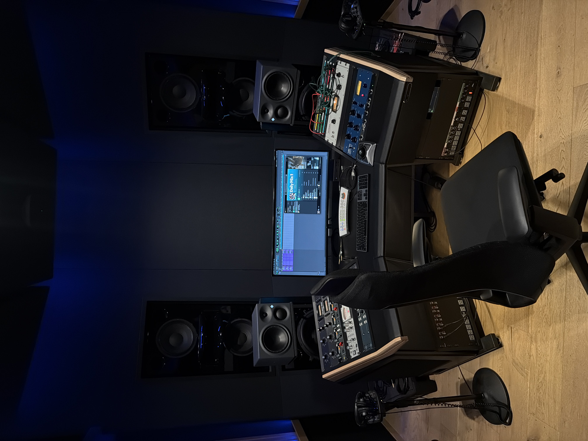 A professional audio studio control room is depicted, designed for mixing or mastering. The room features a dark, acoustically treated environment with a hardwood floor and subtle blue accent lighting originating from the rear wall.
At the center of the setup is a large computer monitor displaying a Digital Audio Workstation (DAW), likely Ableton Live, with various tracks, mixer controls, and an arrangement view visible. A music streaming interface is also open on the screen, showing "Daily Mix 1" and album art. Text visible on the screen includes "M", "Mo", "10", "17:3", "201", "1001", "993", and "00".
Flanking the computer monitor are two large studio monitor speakers integrated into the wall, on either side. In front of these are two smaller near-field studio monitors. On both the left and right of the workstation, slanted equipment racks house a variety of audio processing units, preamplifiers, and interfaces, interconnected by numerous colorful cables. The lower sections of these racks reveal power distribution units or patch bays.
In the foreground, a black office chair is positioned in front of the workstation. To the right of the chair, a microphone stand equipped with a pop filter is visible, with headphones hanging from it. Additional headphones are placed on top of one of the equipment racks. Keyboards and a mouse are present on the desk surface in front of the monitor. The overall atmosphere is dark, focused, and professional, indicative of an environment dedicated to audio production.