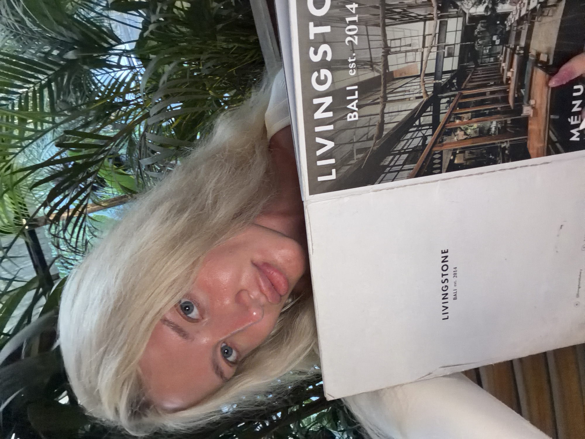 A woman with blonde hair and blue eyes is holding a menu from Livingstone, Bali, est. 2014. She is seated outdoors in a tropical environment, possibly a restaurant, with lush green foliage in the background. The menu features an image of the interior of the establishment, showing tables and chairs. The woman is looking towards the camera, and her face is illuminated by natural light. Her lips are slightly parted, and she appears to be smiling. The overall impression is one of a relaxed and pleasant dining experience.
