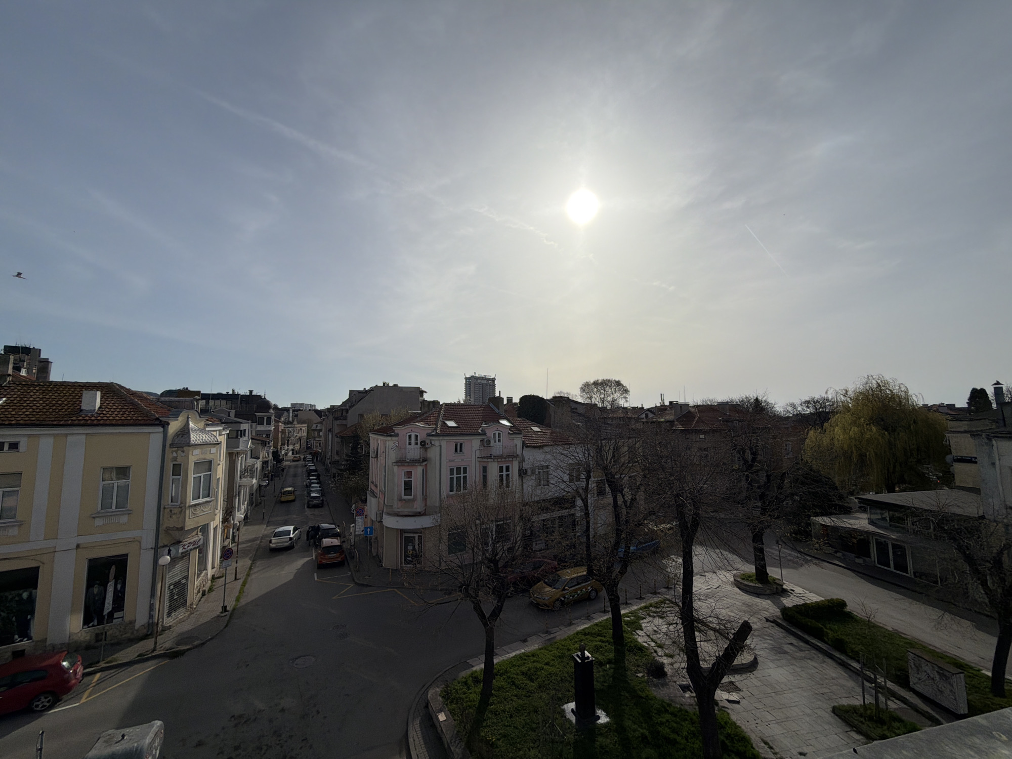 The image depicts an urban scene in Varna, Bulgaria on a sunny day. The view is from an elevated position, overlooking a street intersection and adjacent buildings. The architecture is characterized by older, multi-story buildings with distinct facades, some with balconies and shopfronts at ground level. A line of parked cars is visible along the street. A small park area occupies the foreground to the right, featuring bare trees and paved pathways. In the distance, a taller building stands among the other structures. A bird is flying in the upper left portion of the sky.