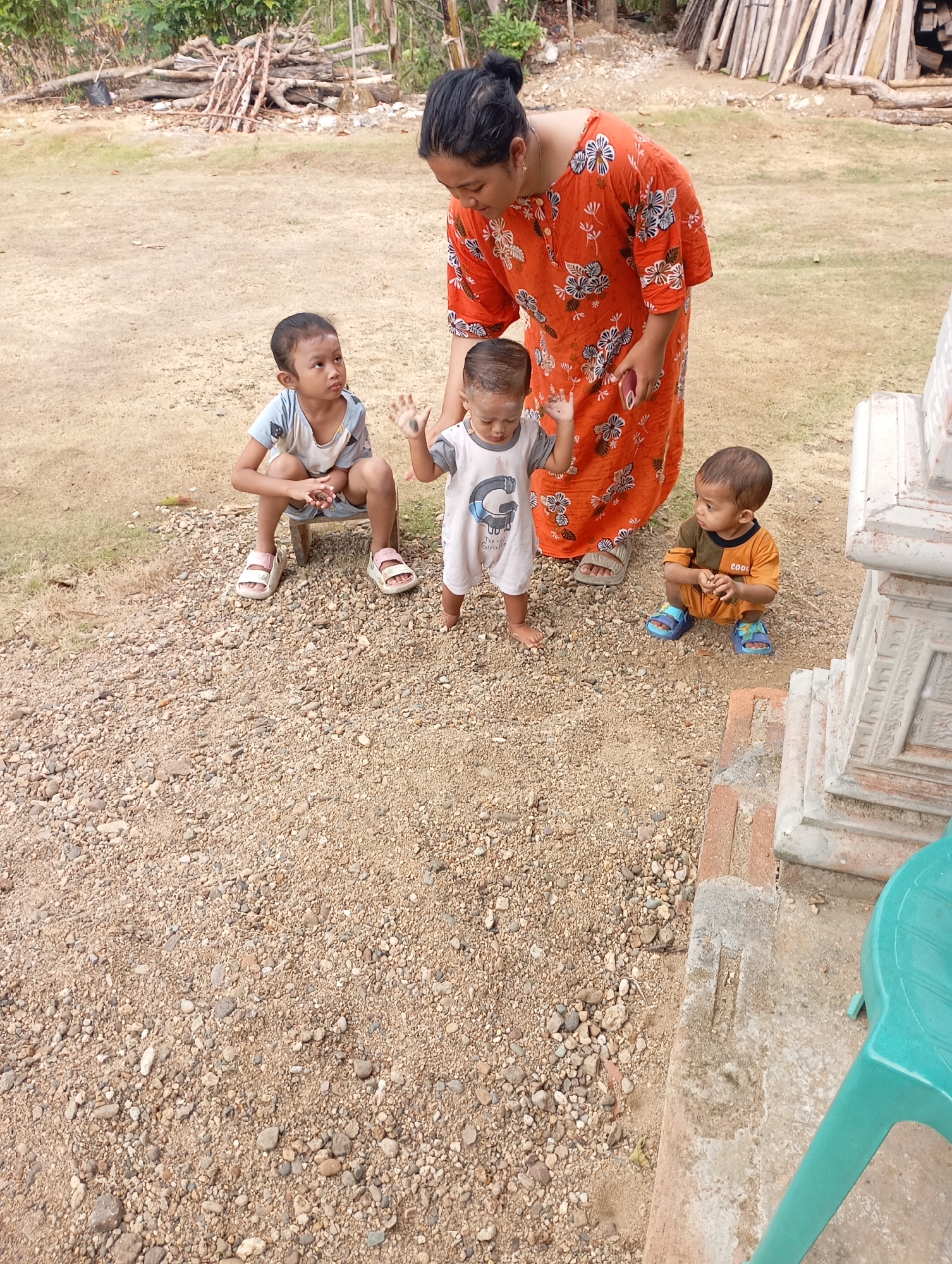 In Ketepung, Indonesia, a woman in an orange floral dress is interacting with three young children outdoors. One child, wearing a light blue and white shirt and shorts, is crouched on the ground. Another child, in a white outfit with gray trim, is standing and has their hands raised. A third child, wearing an orange shirt and shorts, is crouched down. The ground is covered in gravel and dirt, with some dry grass visible. In the background, a pile of wood is stacked, and lush green foliage can be seen. To the right of the frame, a portion of a white structure, possibly a pillar, and a teal plastic chair are present. The scene appears to be a casual outdoor gathering.