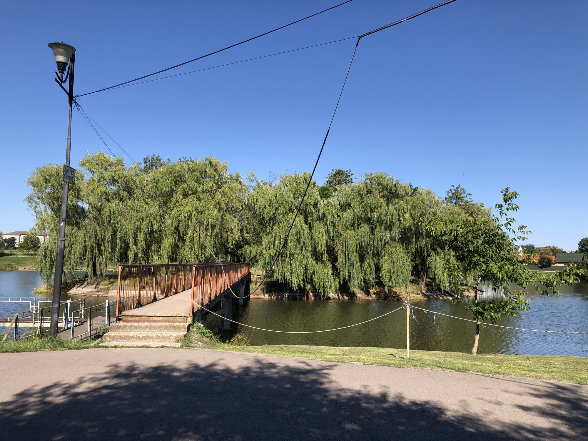 The image depicts a tranquil outdoor scene on a clear, sunny day, likely in the morning or early afternoon given the bright light and shadows. The setting is a park or recreational area bordering a calm body of water, possibly a lake or pond. Dominating the background is a dense line of weeping willow trees, their branches cascading down towards the water's surface, creating a lush, green canopy.
In the foreground, a paved pathway, with distinct tree shadows cast upon it, leads towards a small footbridge. The bridge, constructed with a rusty metal frame and a concrete ramp, spans a section of the water. Concrete steps ascend to the bridge on the right side of the image. A lamppost, tall and dark, stands to the left of the pathway, its light fixture visible against the clear blue sky.
To the left of the bridge, a metal walkway or pier with railings extends into the water, suggesting a point of access for boats or a viewing platform. A simple rope barrier, supported by wooden posts, runs along the edge of the grassy bank, separating the path from the water.
In the distance, across the water and beyond the trees, glimpses of residential buildings with pitched roofs can be seen, hinting at the proximity of a town or village. The overall atmosphere is one of peacefulness and natural beauty, inviting recreation and relaxation. No people are visible in the frame. There is a partially obscured sign attached to the lamppost, but the text is illegible.