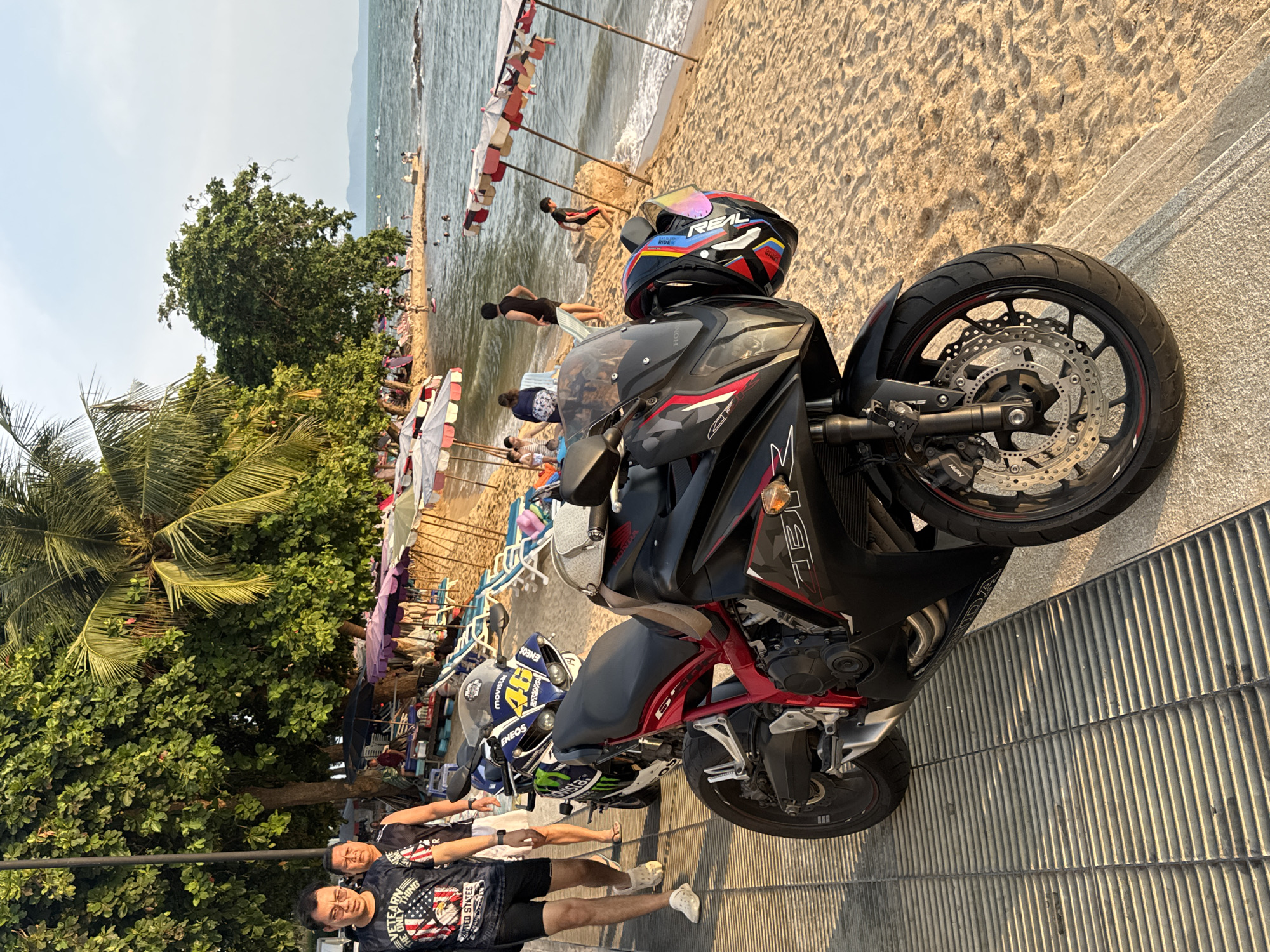 The image depicts a black and red Honda motorcycle parked on a concrete walkway in Pattaya City, Thailand. A helmet rests on the motorcycle. In the background, a beach scene unfolds with people relaxing under colorful umbrellas. Two men are standing near the motorcycle on the walkway. The scene is bathed in natural sunlight, suggesting it's daytime.