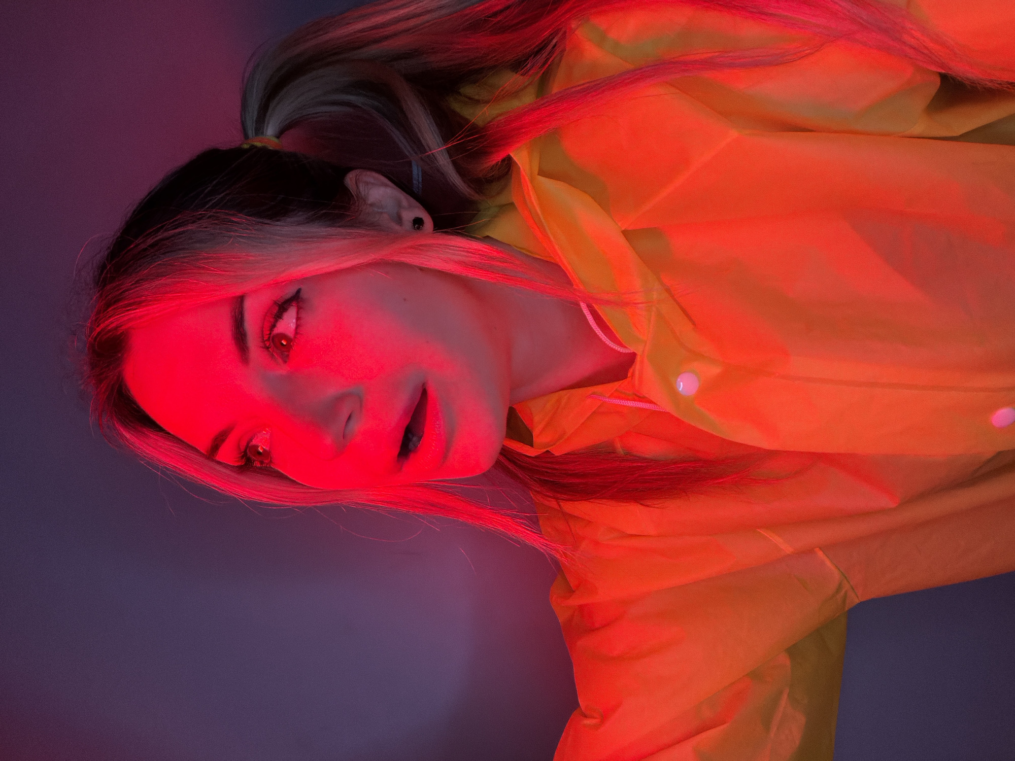 A vertically oriented, stylized portrait captures a young person, likely female, illuminated predominantly by an intense red light. The subject's head is tilted sharply to the left, with their eyes wide and directed upwards, and lips slightly parted, showing dark lipstick. Their face is bathed in a strong red glow, highlighting dramatic eye makeup, including winged eyeliner. A small black stud earring is visible in their left ear.
The person has light-colored hair with darker roots, pulled back into a ponytail with a yellowish hair tie. The red light makes sections of their hair appear bright crimson. They are wearing a vibrant yellow or orange raincoat or jacket, which also reflects the dominant red lighting, making it appear reddish-orange. Visible details of the jacket include snap buttons and subtle fabric folds.
The background is a plain, dark surface, transitioning from a deep purple-blue at the top to darker tones at the bottom, providing a stark contrast to the brightly lit subject. The powerful red illumination creates a high-contrast, dramatic, and somewhat mysterious atmosphere. No specific activity beyond posing for the photograph is evident. There is no visible text in the image. While the location is provided as Budva, Montenegro, the visual content does not offer specific cues to confirm this geographical setting.